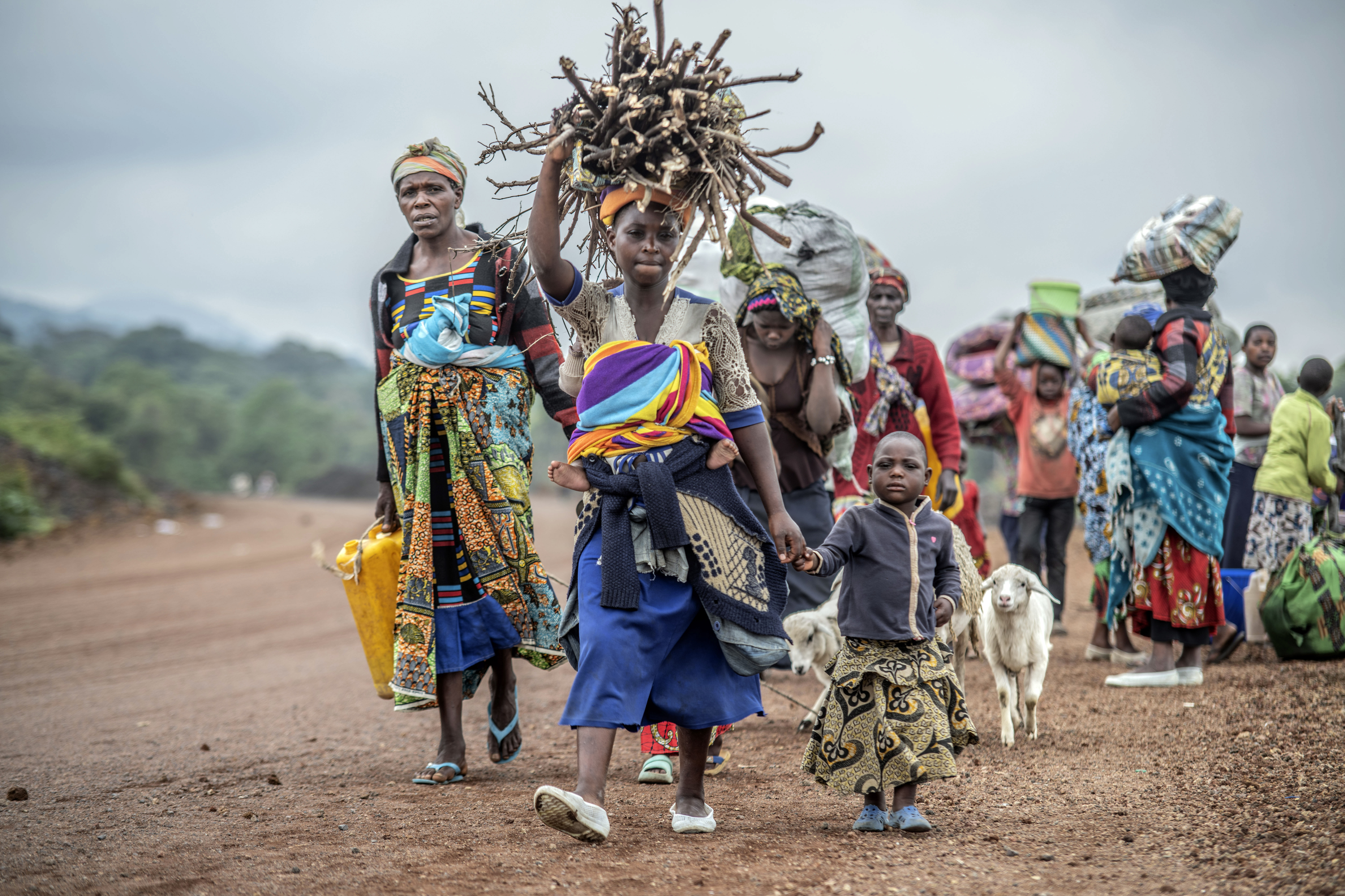 Women wearing colourful clothes, one carrying a yellow petrol canister and others bags of food on their head, flee fighting between M23 rebels and Congolese forces . There are also goats and children in the line of people.
