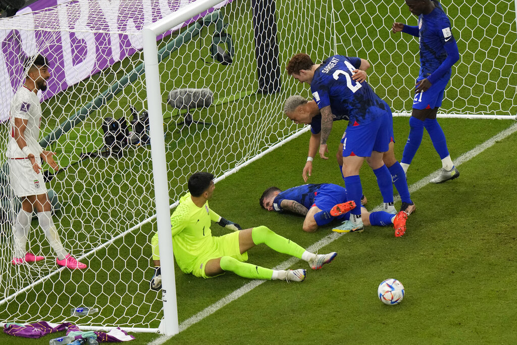 Pulisic lying on his stomach between the goal posts, face in agony, as the goalie sits looking at him while teammates bend down to check on him