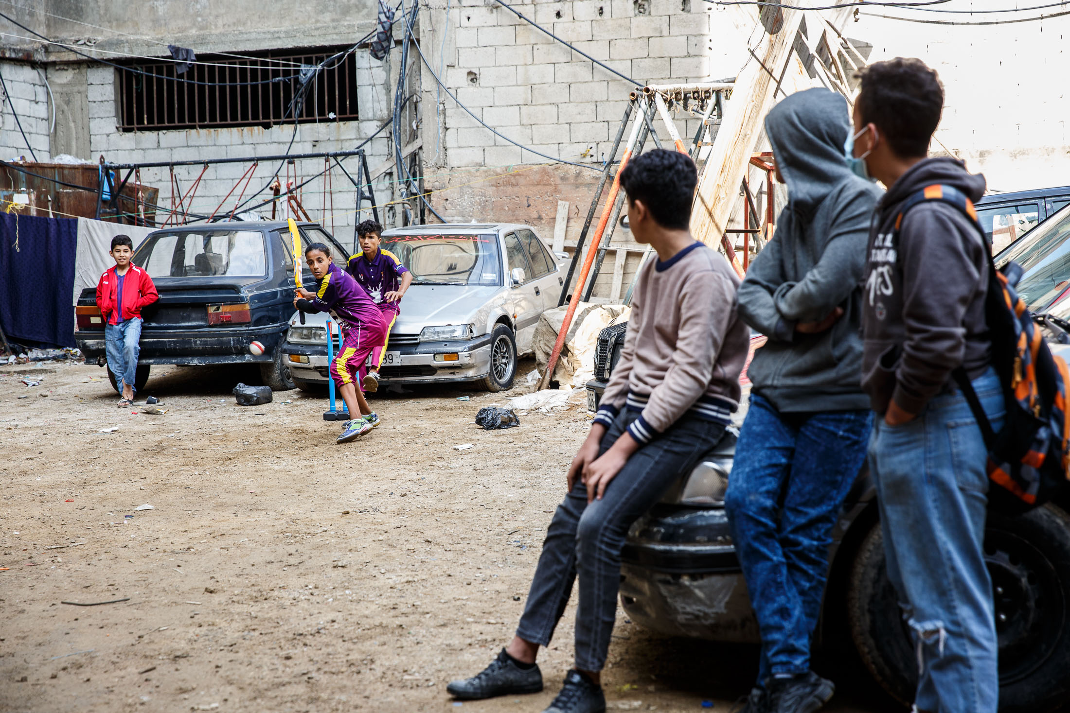 Children sitting on a car watch others play cricket in an urban environment