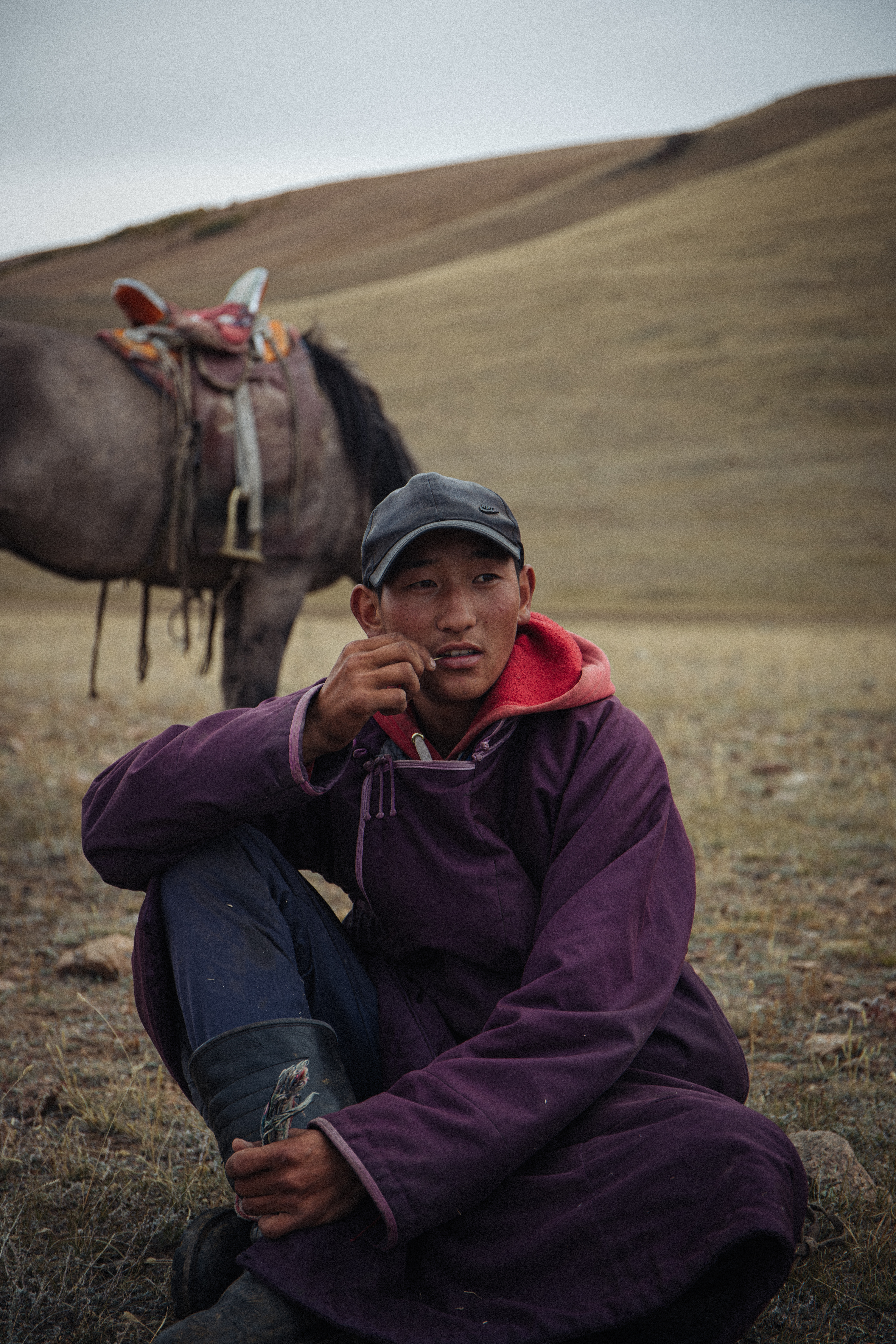 Shwara, 18, hopes to join the Winter Herd one day. He is sitting in the brown grass looking pensive with his horse grazing behind him