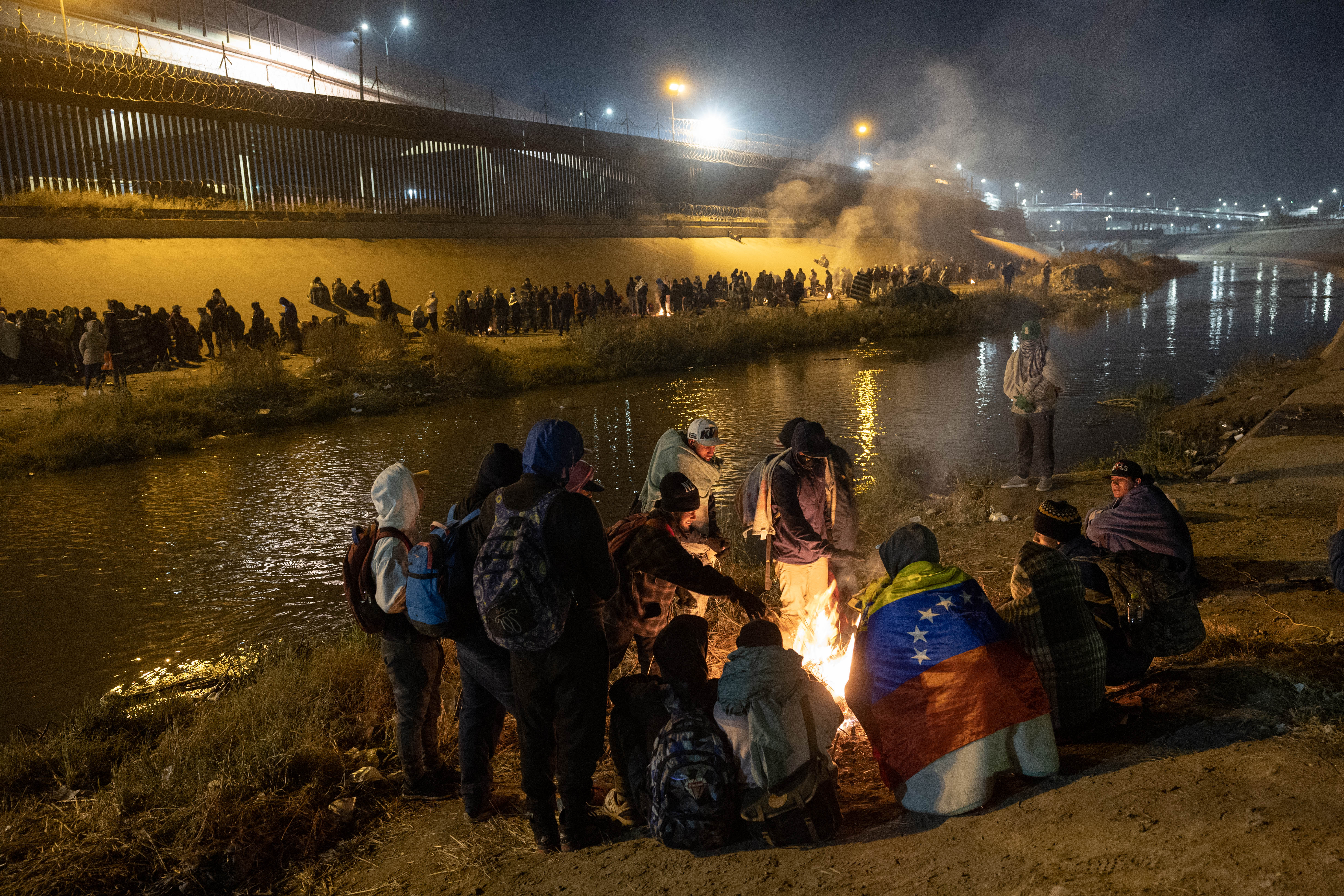 People around fire on a small river bank. On the other side of the river, people line along the whole bank. There are some fires lit there too