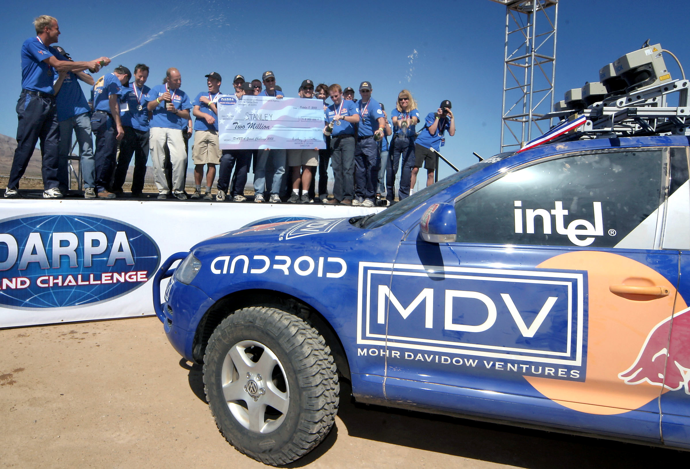The Stanford Racing Team, atop a podium, sprays champagne with their winning autonomous vehicle on the road below