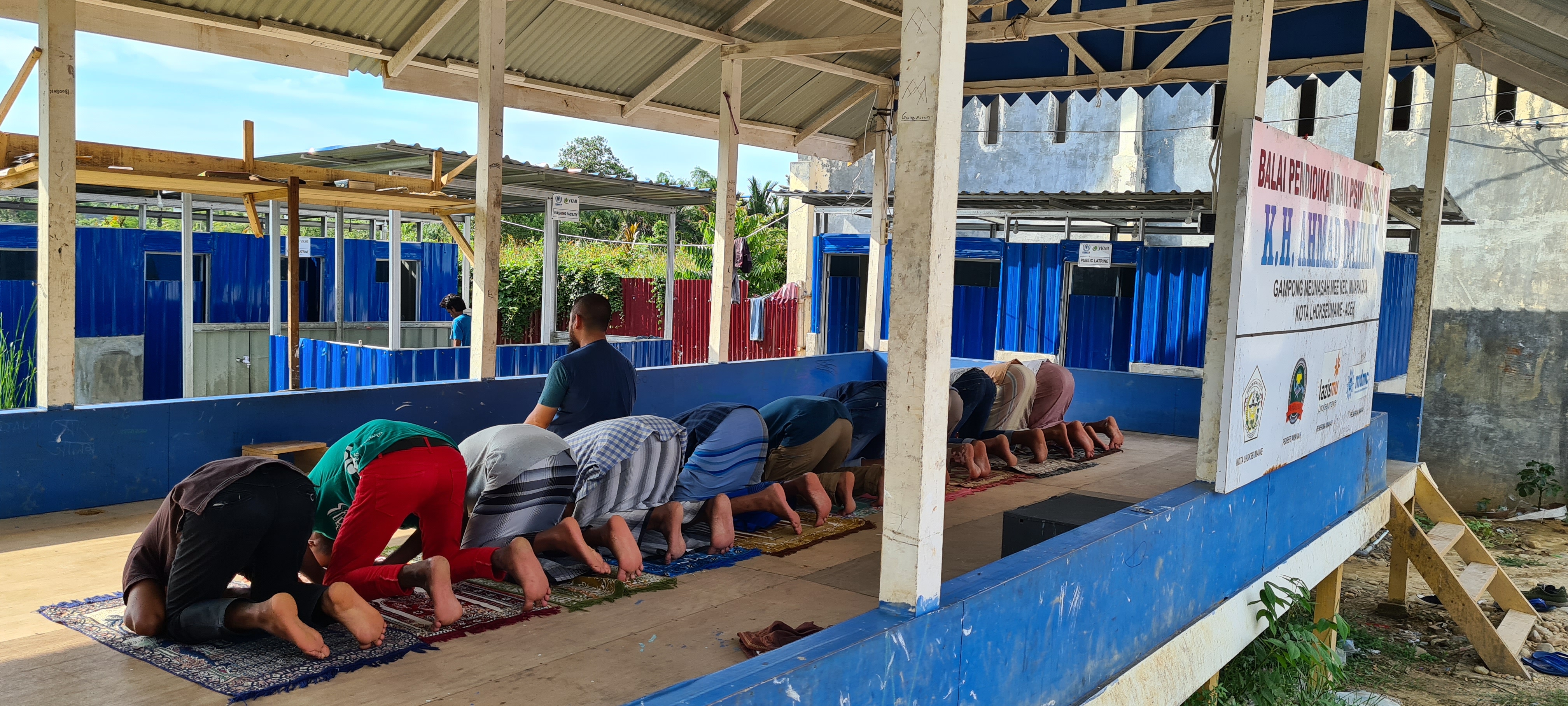 The Rohingya refugees praying at the temporary camp in Aceh. They are beneath a shelter. The newly built toilets are in the background