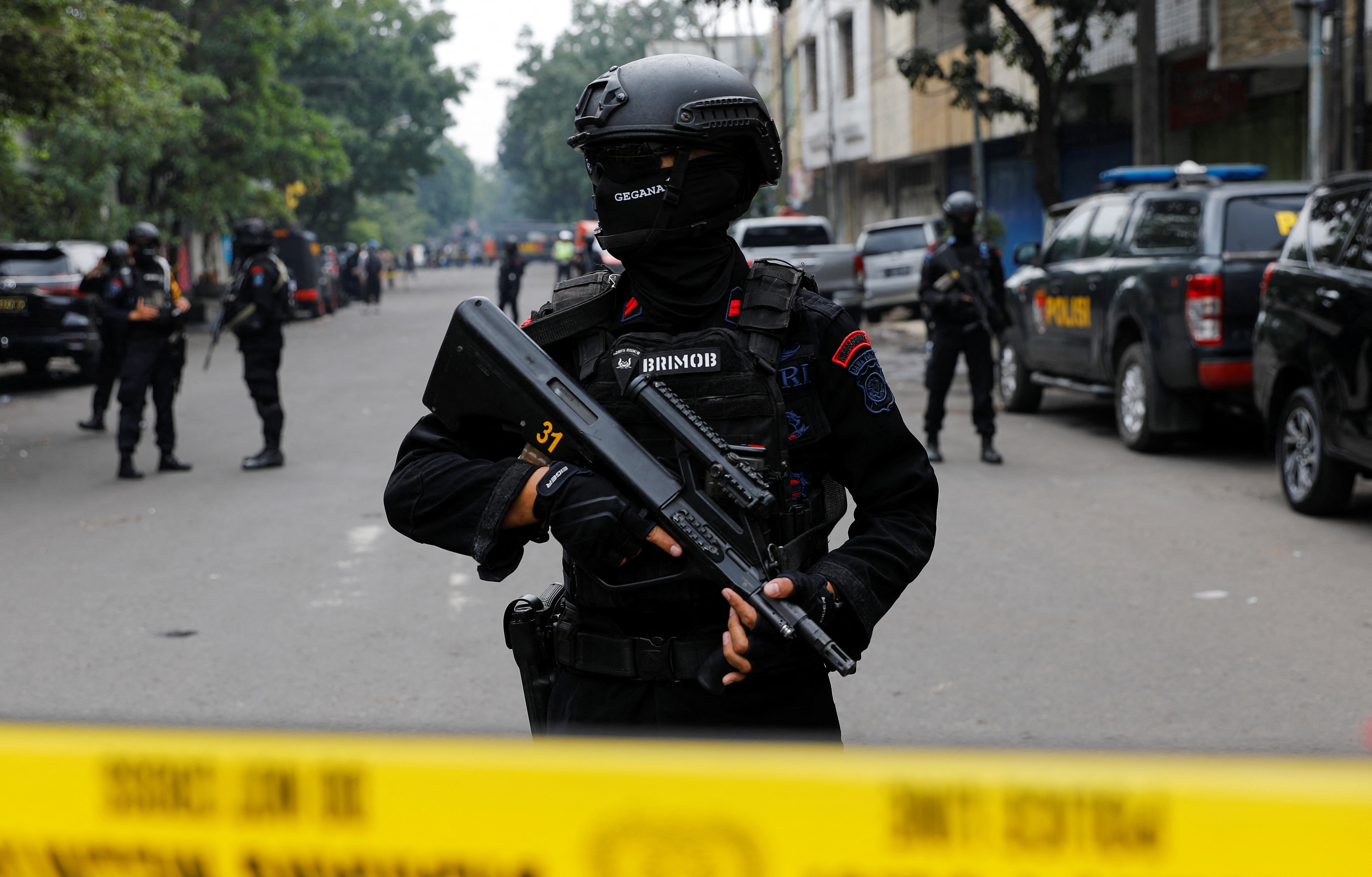An Indonesian police officer, wearing a black BRIMOB uniform and carrying a weapon. Only his eyes are visible. He is carrying a weapon and standing behind yellow police tape