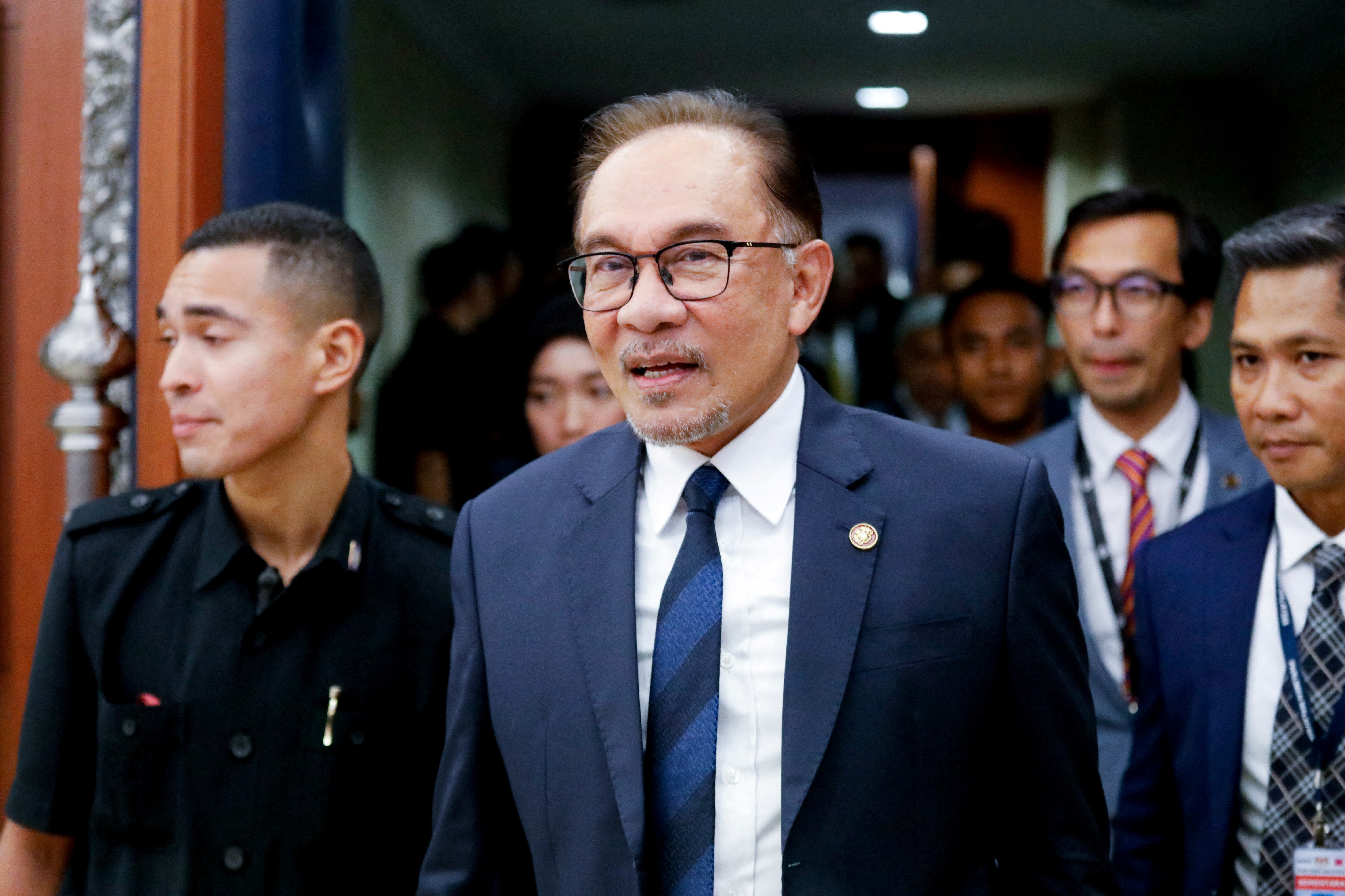Malaysia PM Anwar Ibrahim wearing a blue suit and tie walks out of a hallway with a people behind him and next to him wearing lanyards.