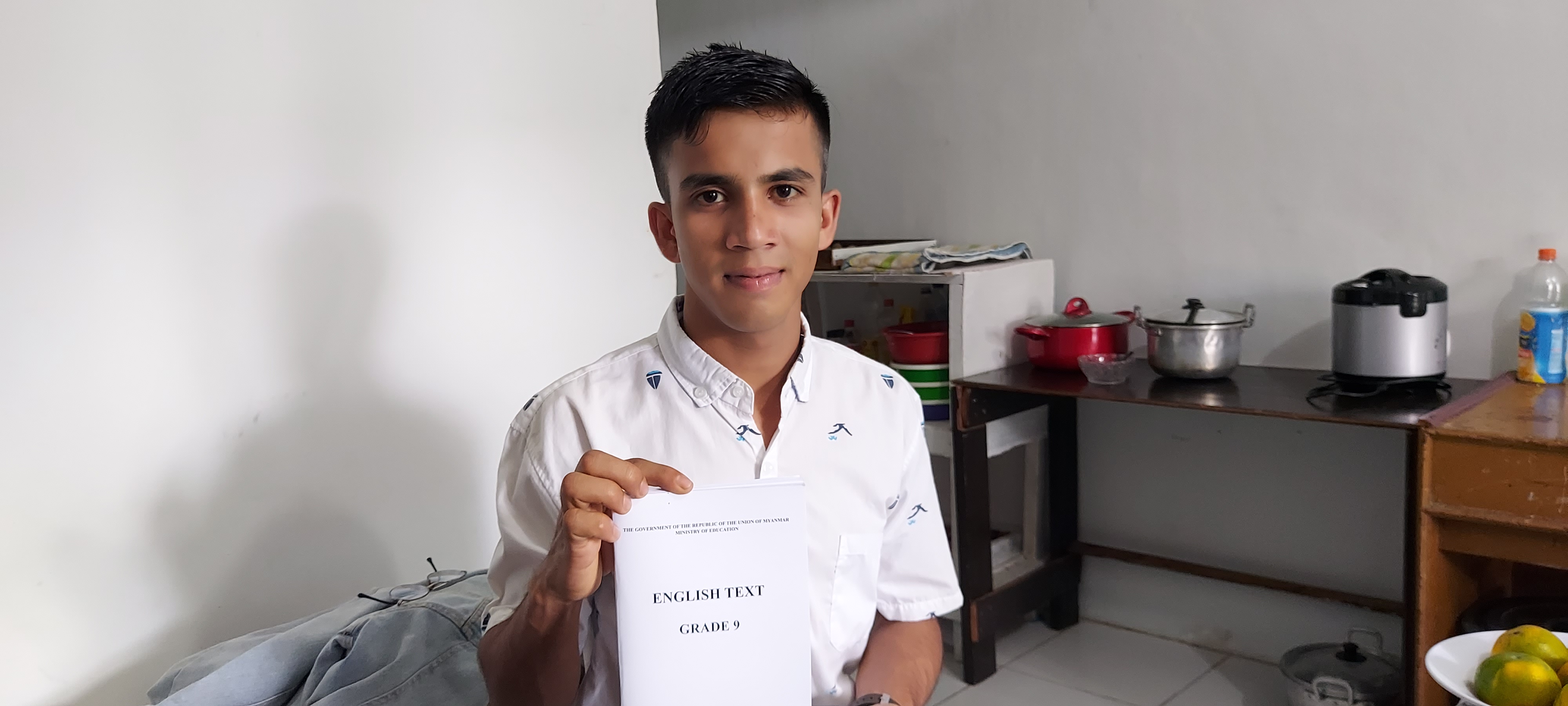 Gura Amin sitting in his room holding up a copy of a new English book 'English Text, Grade 9', He looks pleased. He is sitting on his bed. The walls are bare. There are pots and a rice cooker on the table behind him
