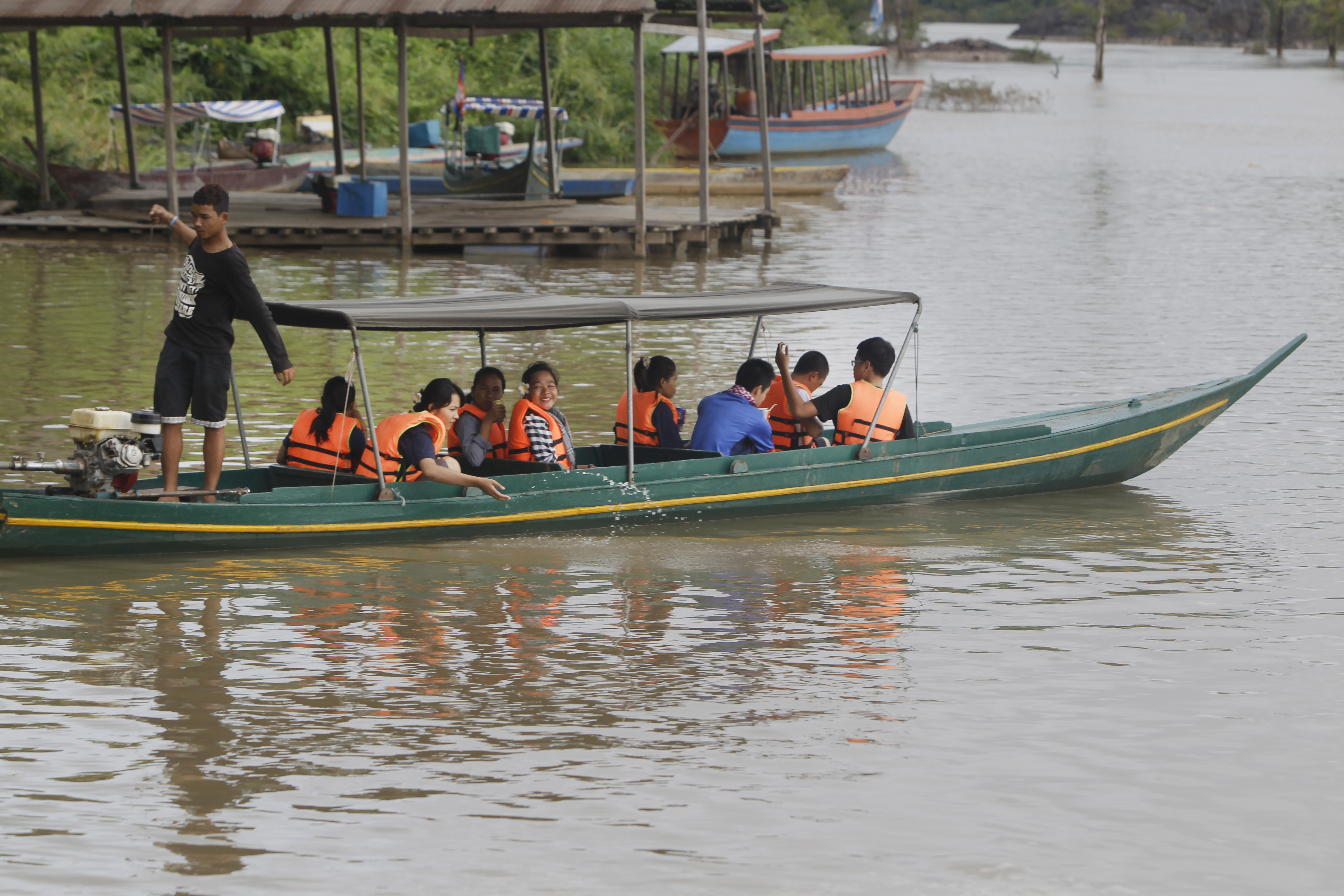 A boat filled with tourists set off onto the Mekong in the hope of catching a glimpse of the Mekong dolphin. The people are wearing life jackets and the boat is uncovered. There are other boats moored along the bank behind them