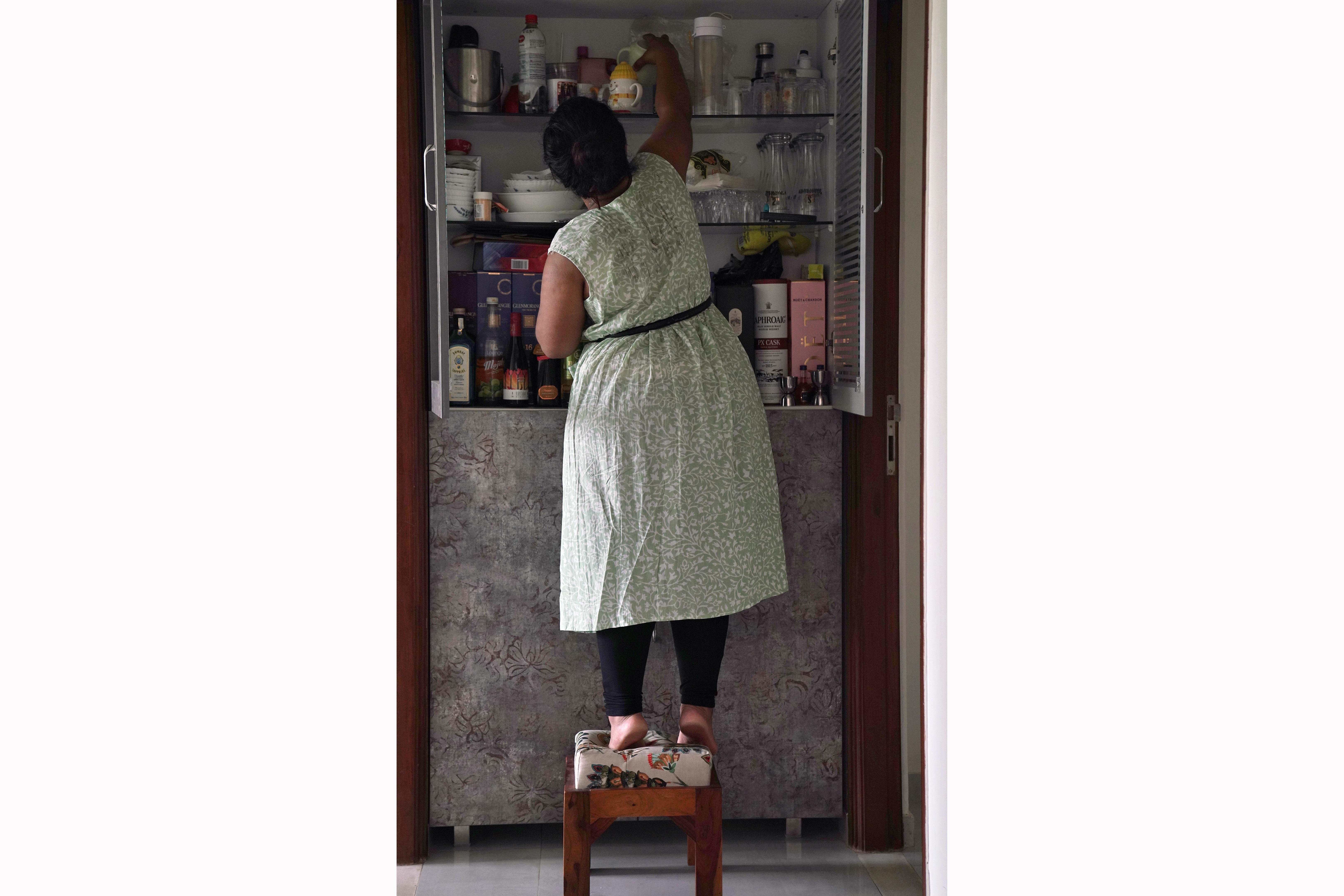 Akanksha stands on a stool in front of her pantry cupboard, lifting onto her toes to reach something in the top shelf