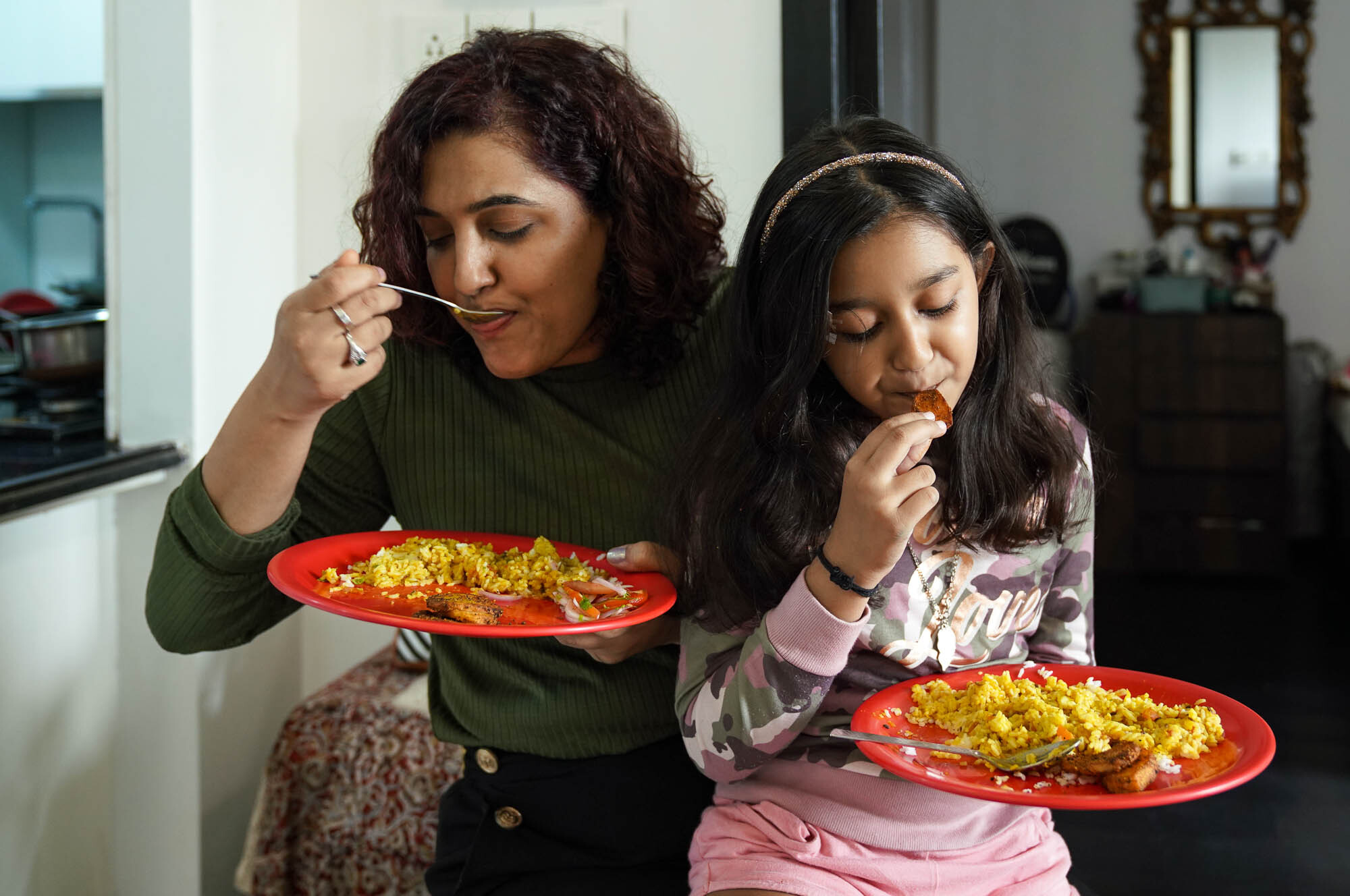 Khushboo sits with her daughter on her lap, each eating their meal out of a red plate