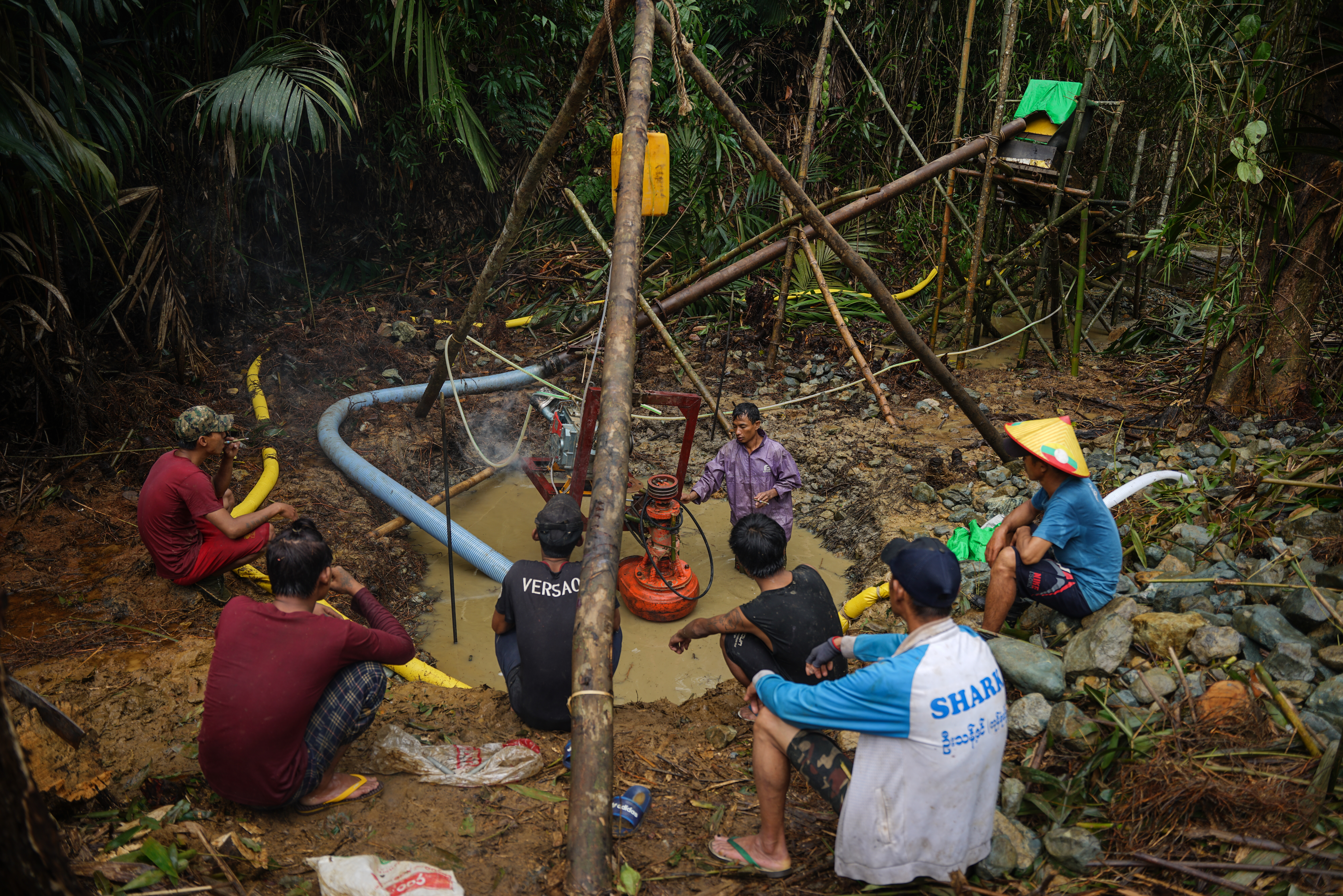 Six men gathered around a mining pool in a clearing cut in the jungle for gold mining. A seventh man is up to his thighs in the water working a machine