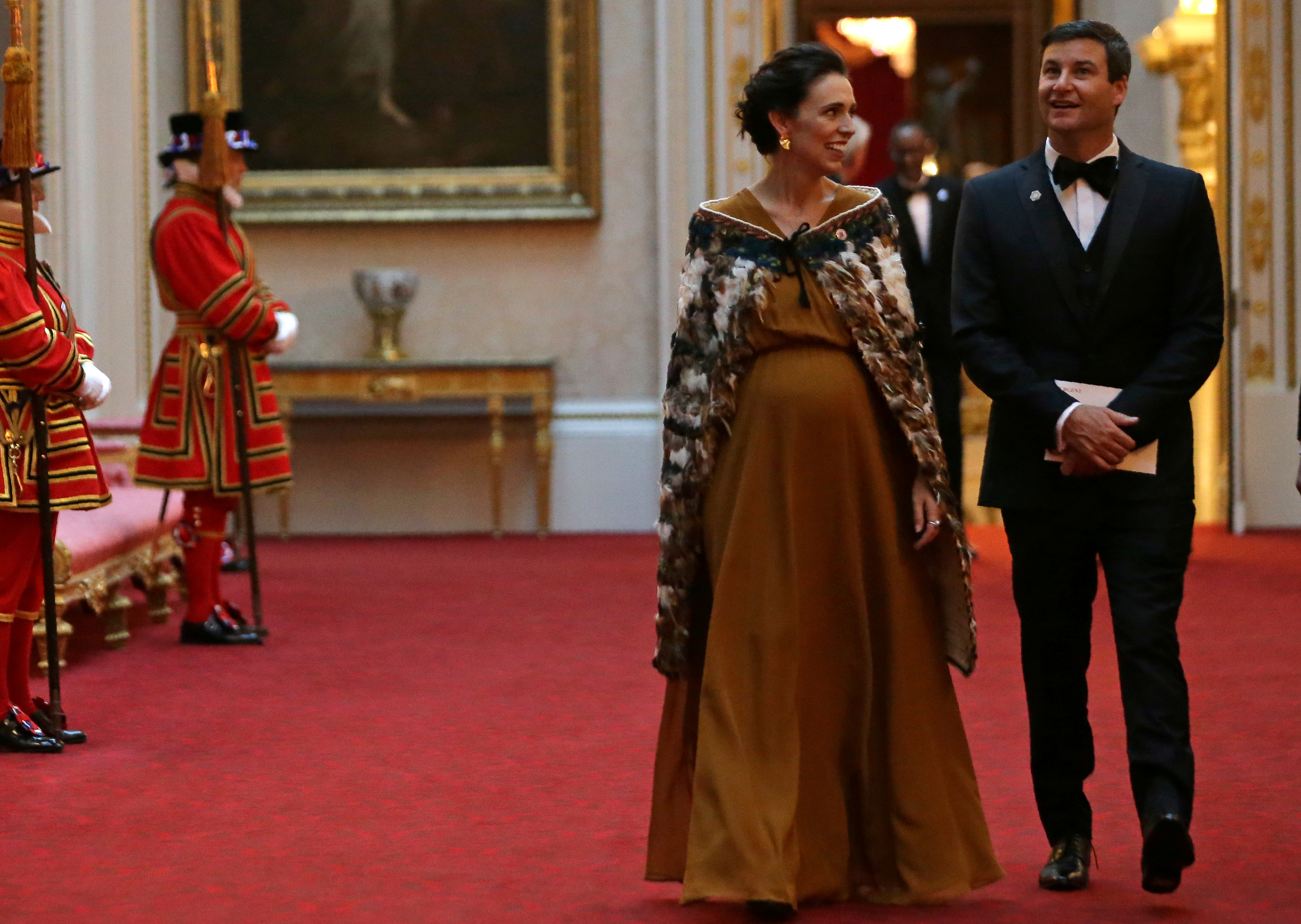 Jacinda Ardern with her partner walks through Buckingham Palace wearing a brown silk dress and a traditional Maori cloak known as a kaxx, They are both smiling. A beefeater is standing to the left and there are paintings and antiques behind them
