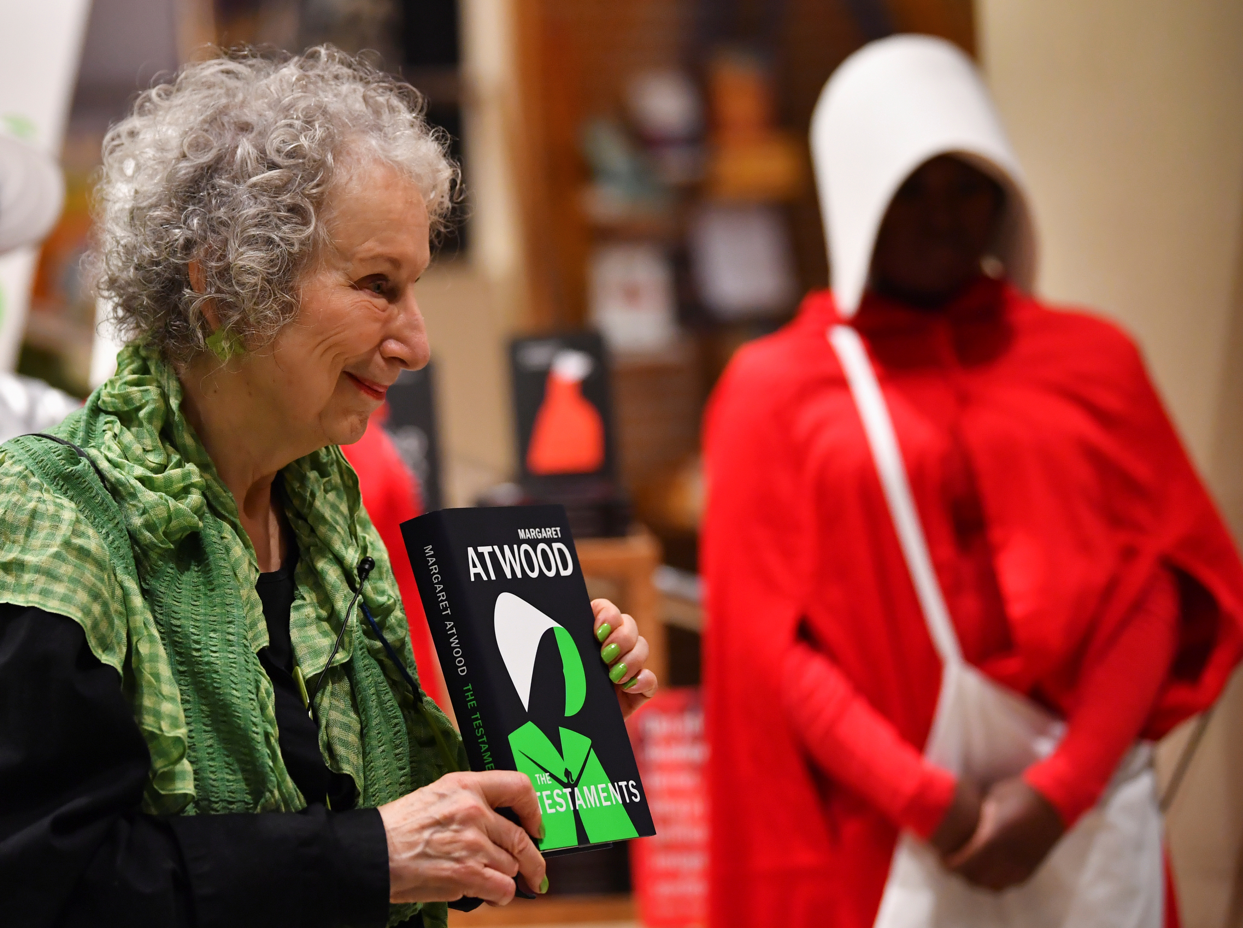 Margaret Atwood holds up a copy of "The Testaments" while an event attendee wears a costume of the handmaids – a red robe and white bonnet that covers the woman's peripheral vision