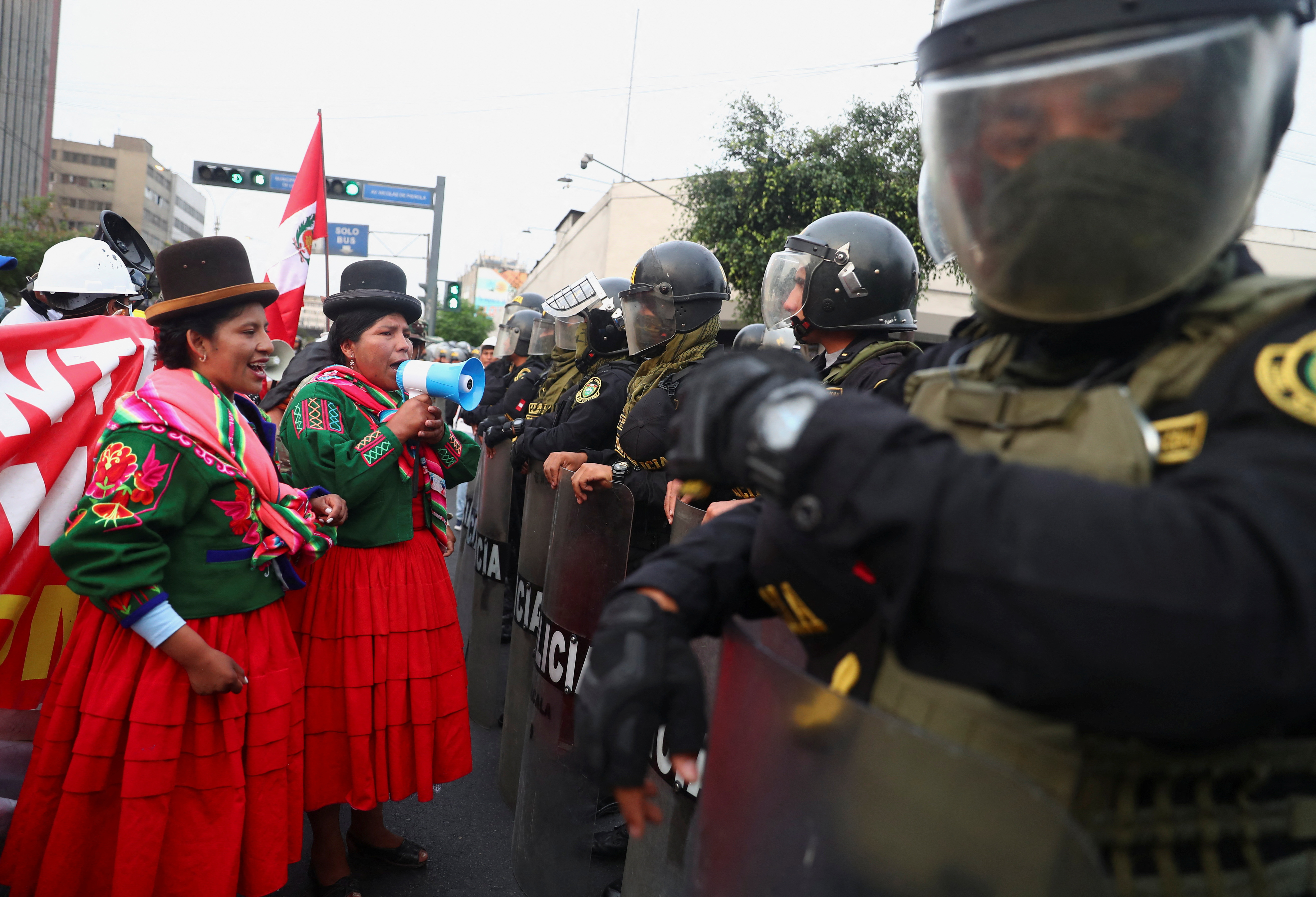 Two women in traditional Peruvian dress face ranks of riot police wearing helmets and carrying shields. One of the women is speaking through a loud hailer