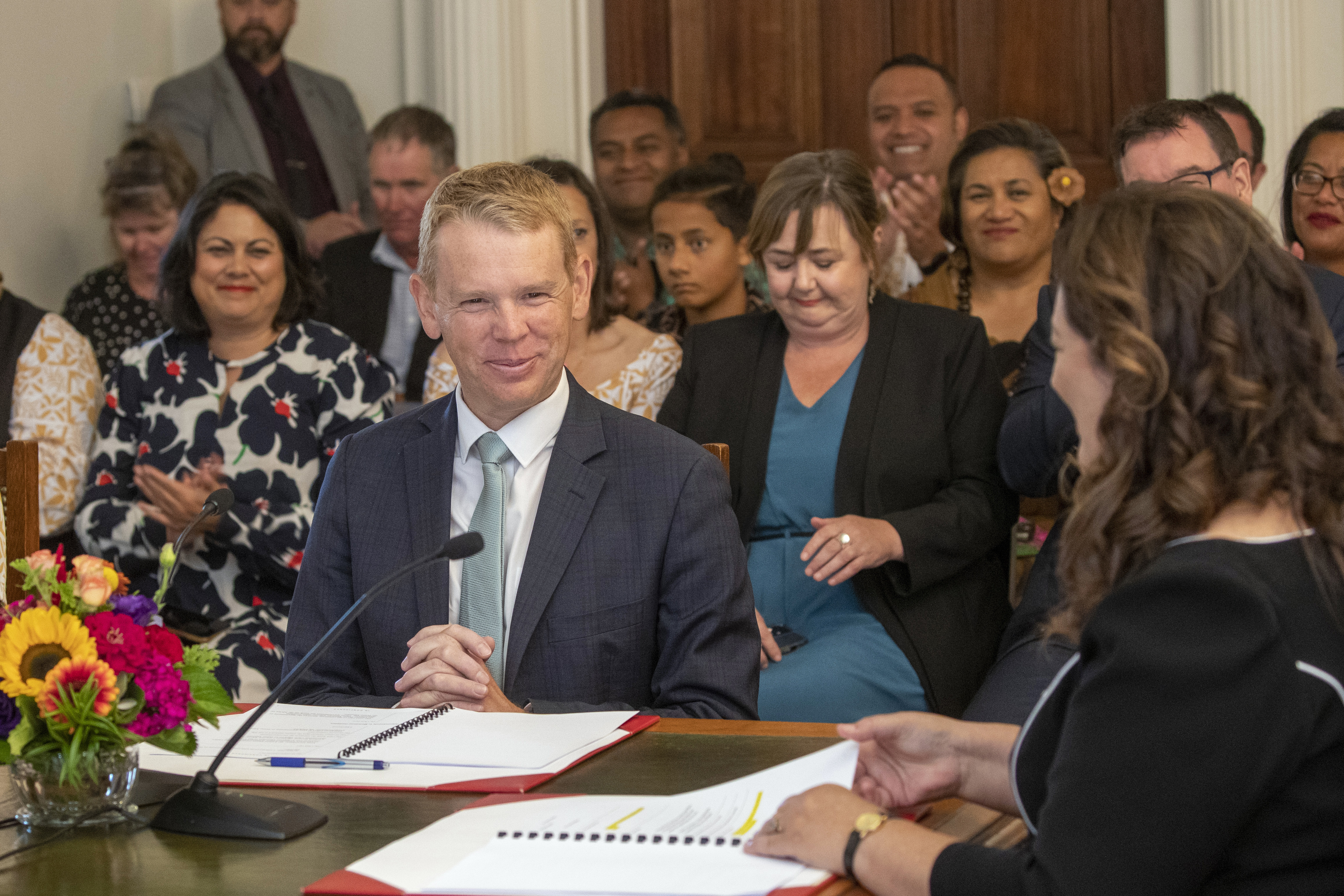 Chris Hipkins is sworn in as Prime Minister. He is sitting at a table with Governor General Cindy Kiro who is on the right of frame. They have papers on the table in front of them and there is a flower arrangement in the centre. There are people watching, most of them are women. They look happy