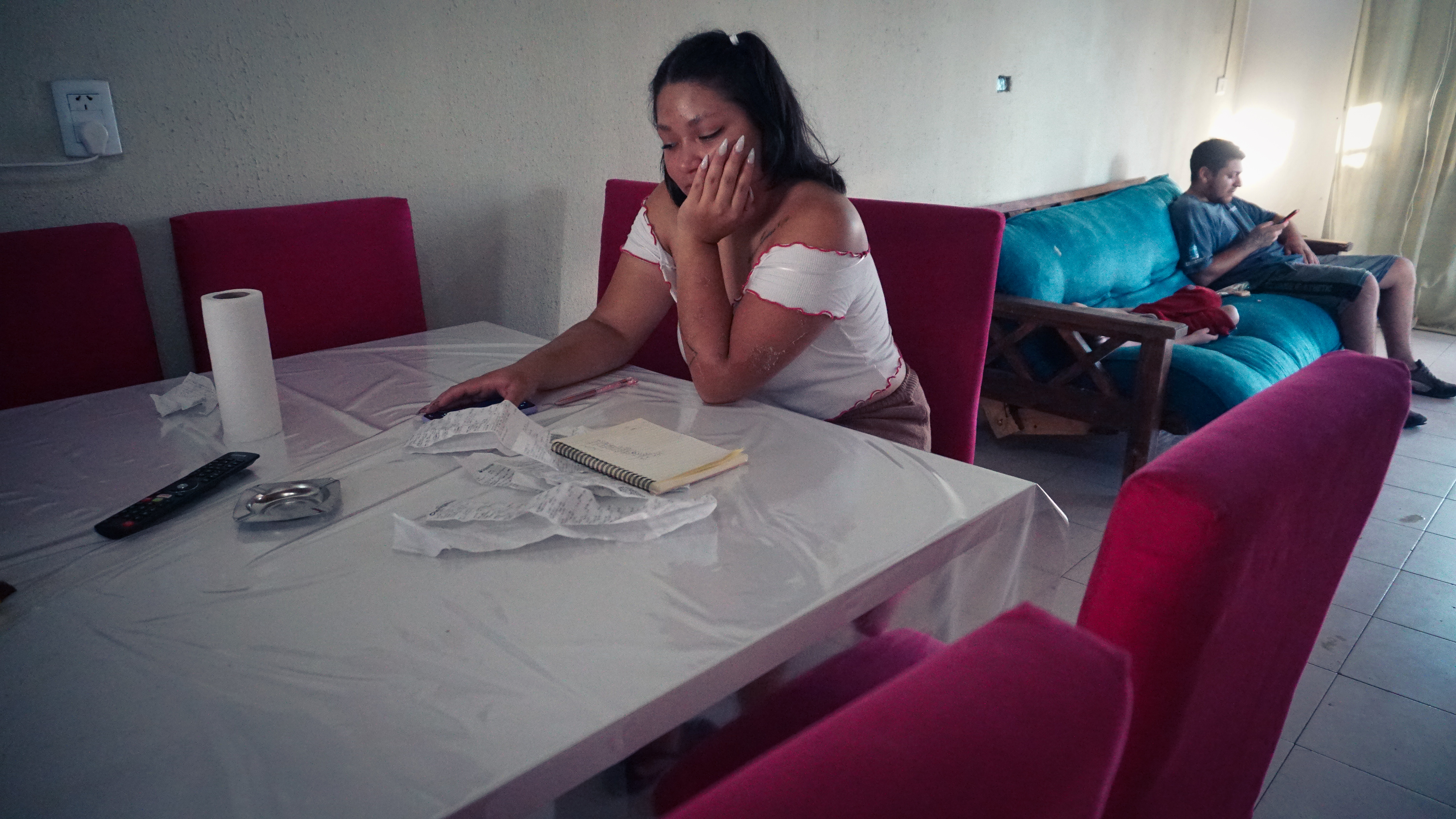 A photo of a woman sitting and looking at some receipts on a table with a man sitting on a sofa behind her.