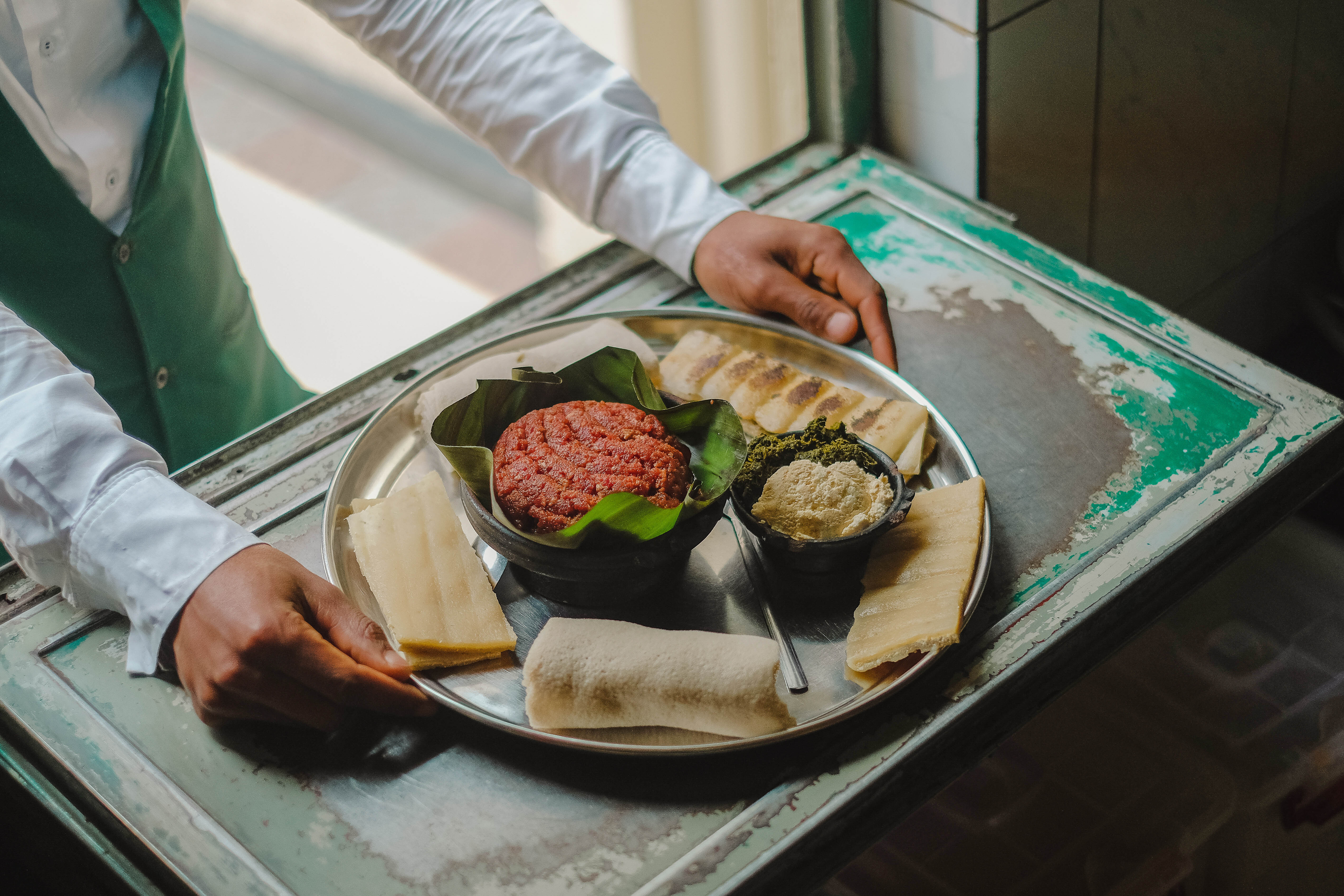 A waiter's hads at the pass taking a prepared kitfo tray from the pass out to a cusomer