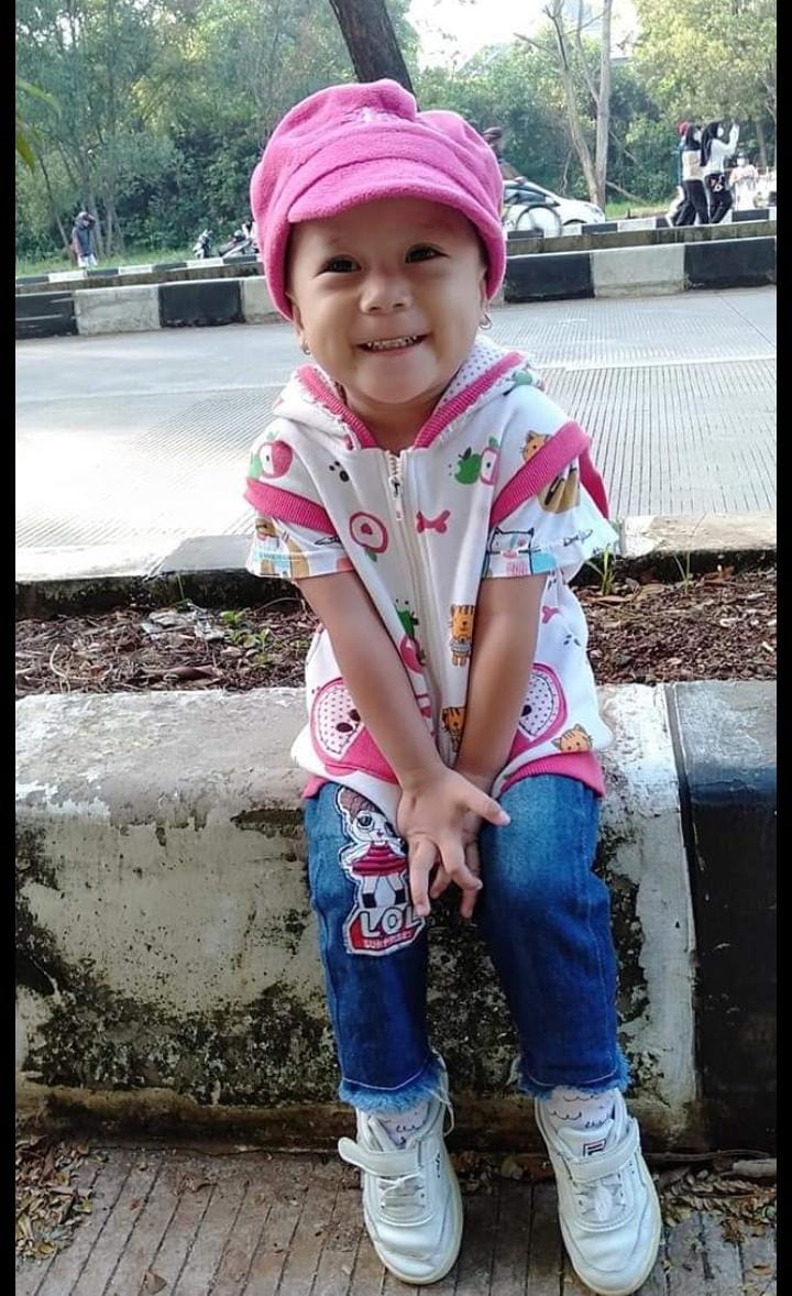 Azqiara sitting on the kerb by the side of the road. She is wearing jeans, a pink and white short and a pink hat. She is smiling and looks really happy