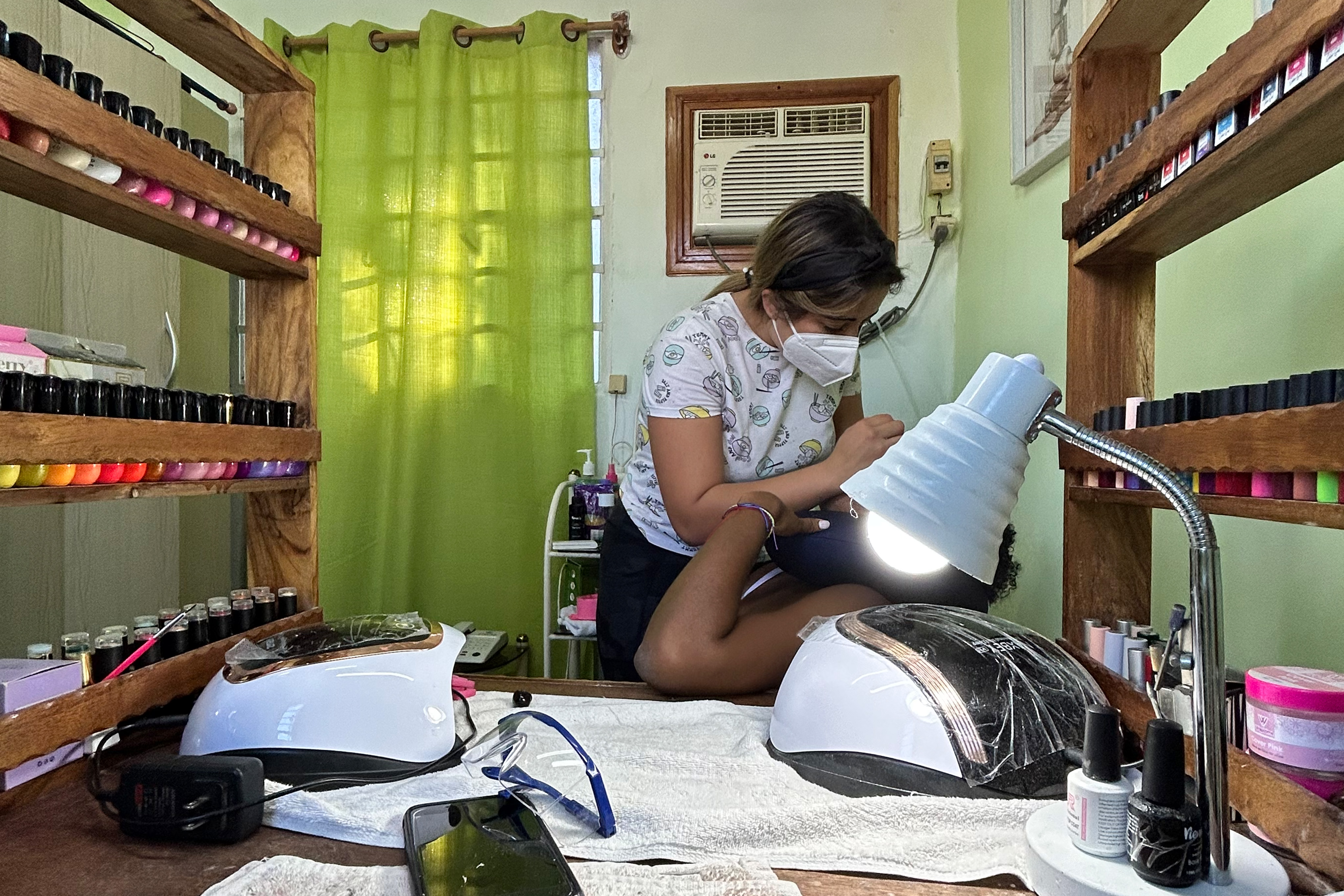Patri at home, applying makeup to a client surrounded by shelves. A desk covered in papers sits in the foreground.