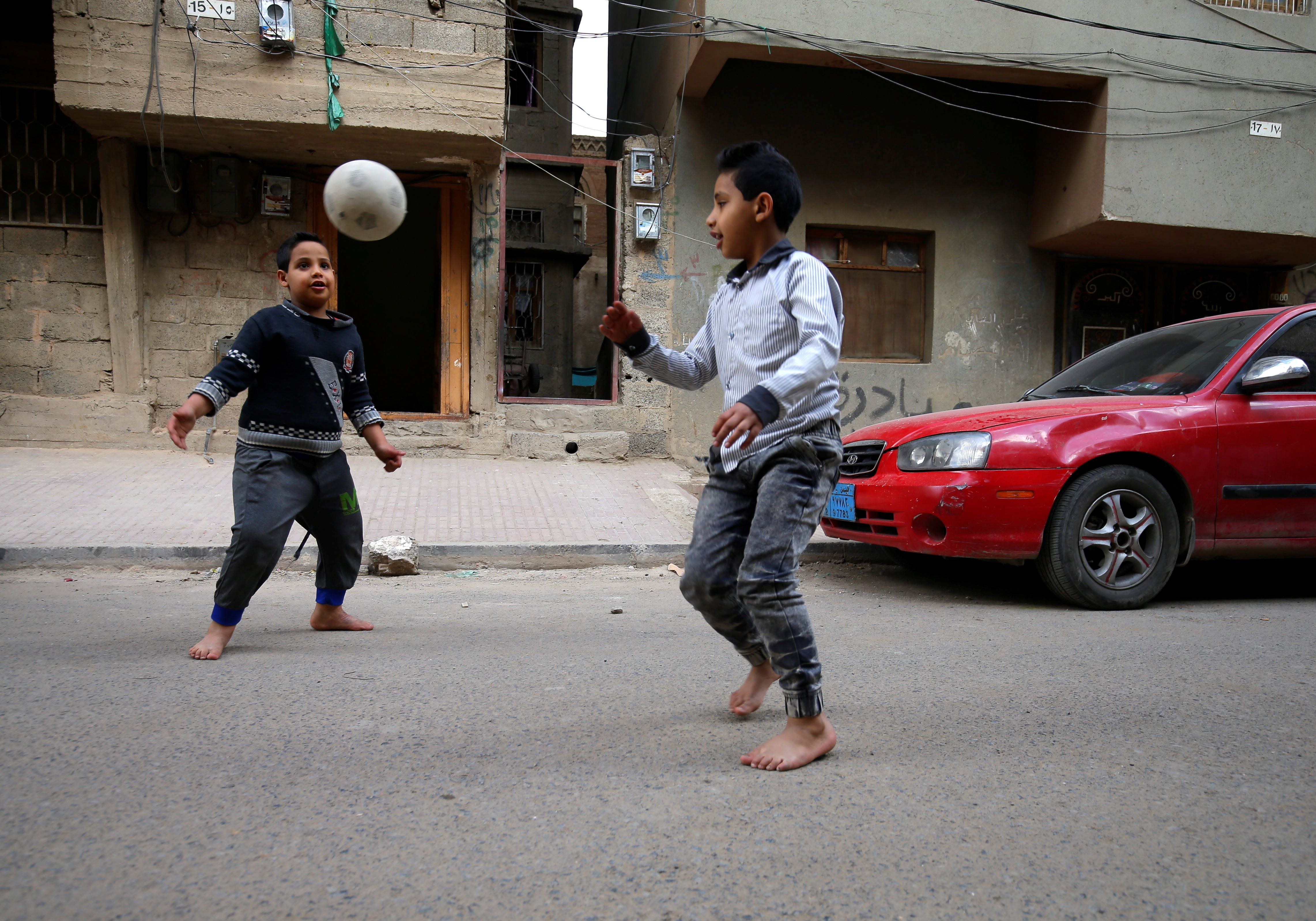 A photo of two kids playing football in the street, outside of a building with a red car in front of it.