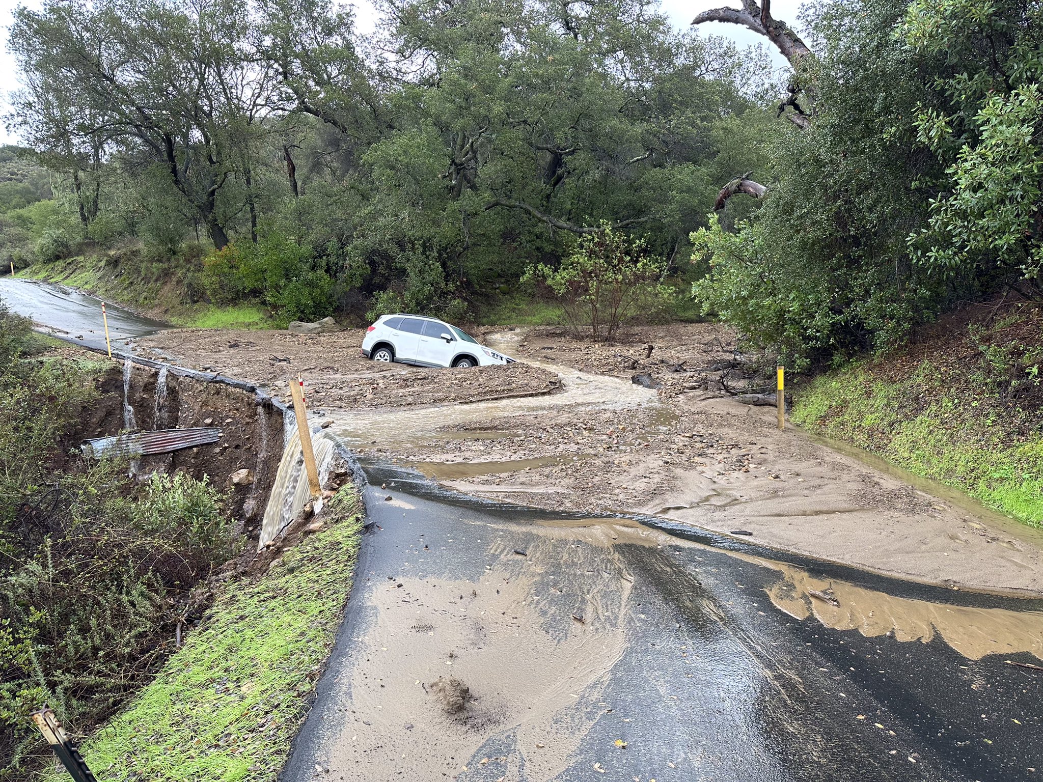 A car lays trapped in mud on a washed out road. There are trees on either side