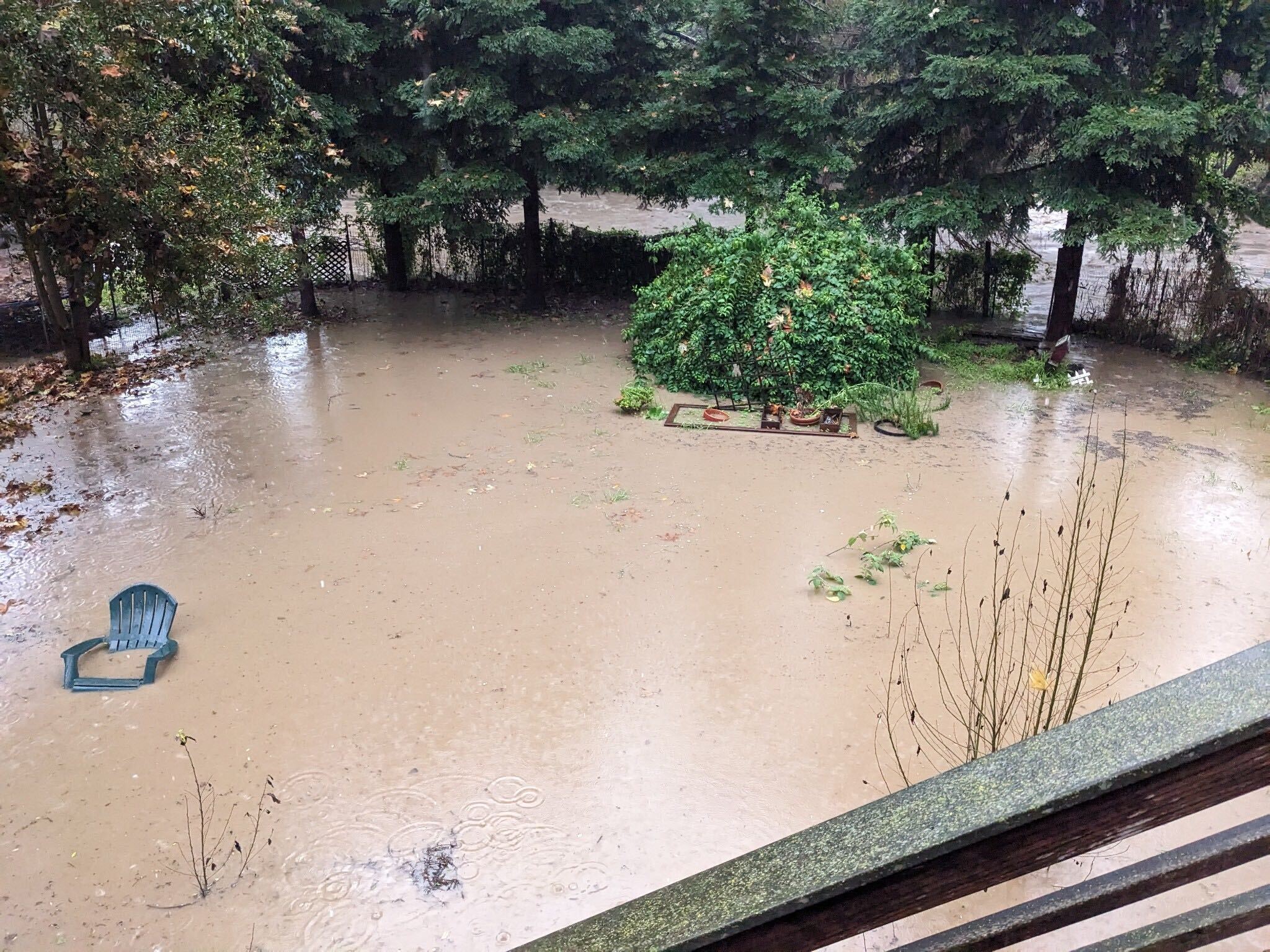 The backyard is full of muddy water. There a plastic chair sitting in it with the water rising up to the side handles.
