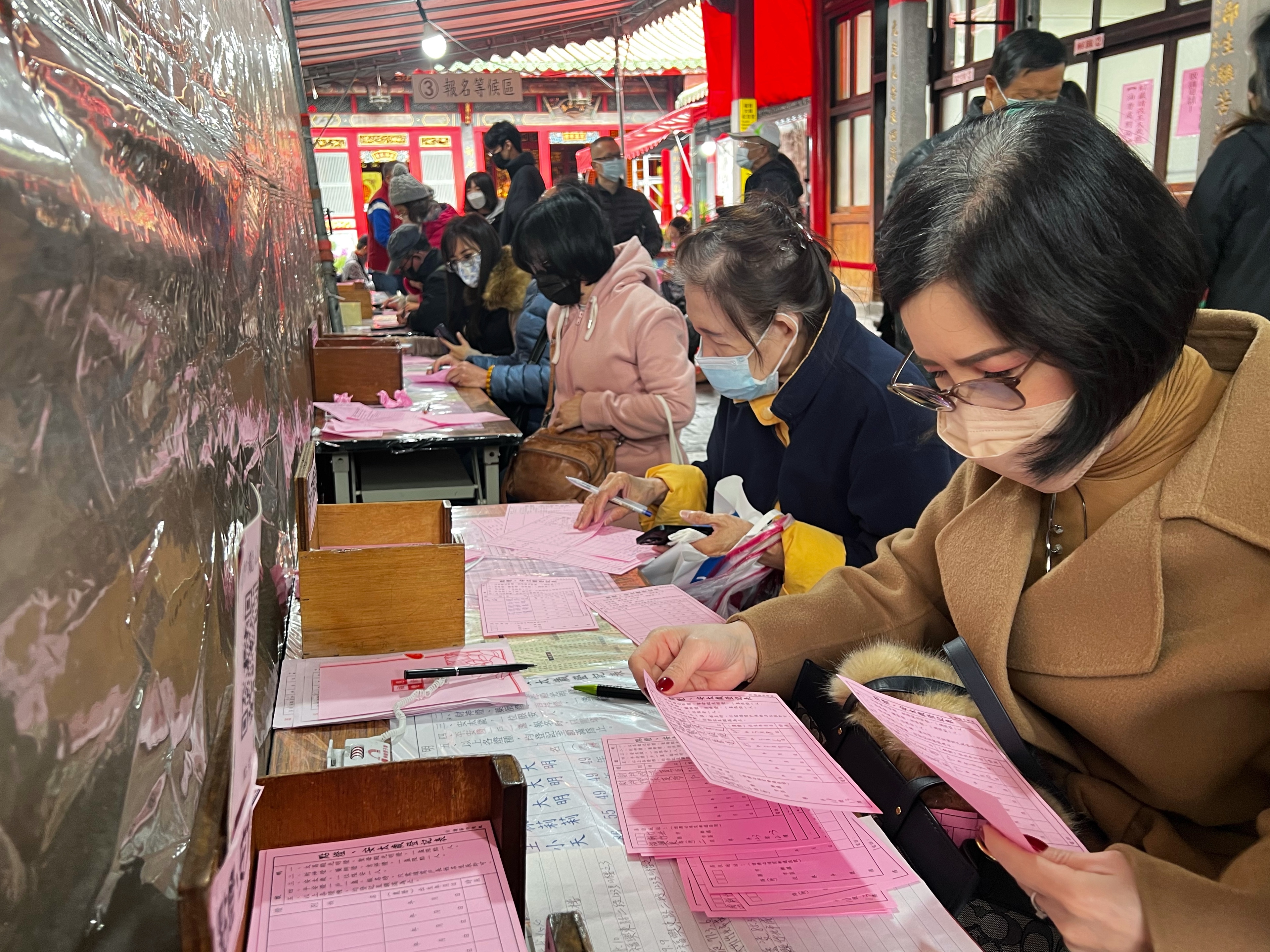 A woman in coat and face mask fills out her particulars so the gods at the Longshan temple will know her. The form is pink and there are lots other people filling it out as well