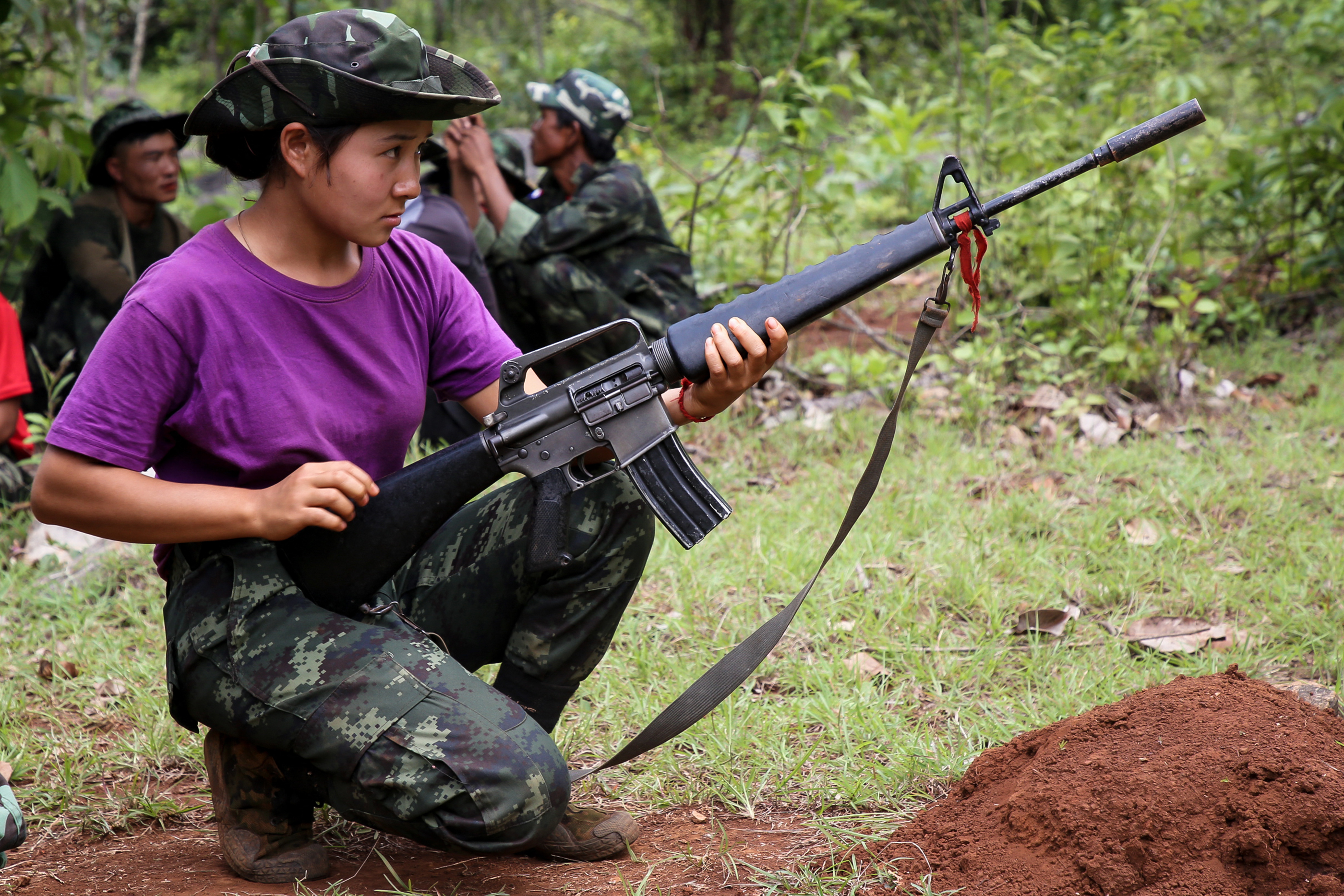 A Karenni woman in a military training session with a weapon. She is wearing a purple t-shirt and fatigues. She looks focused.