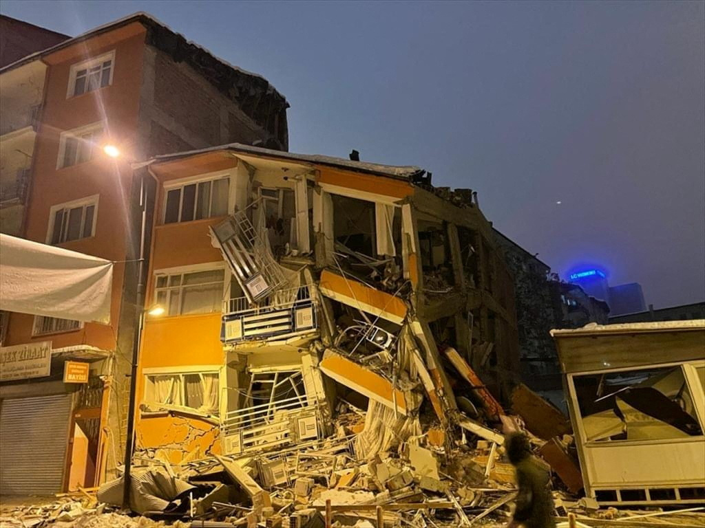 A collapsed building in Malatya, Turkey. The building is illuminated. A man is walking past.