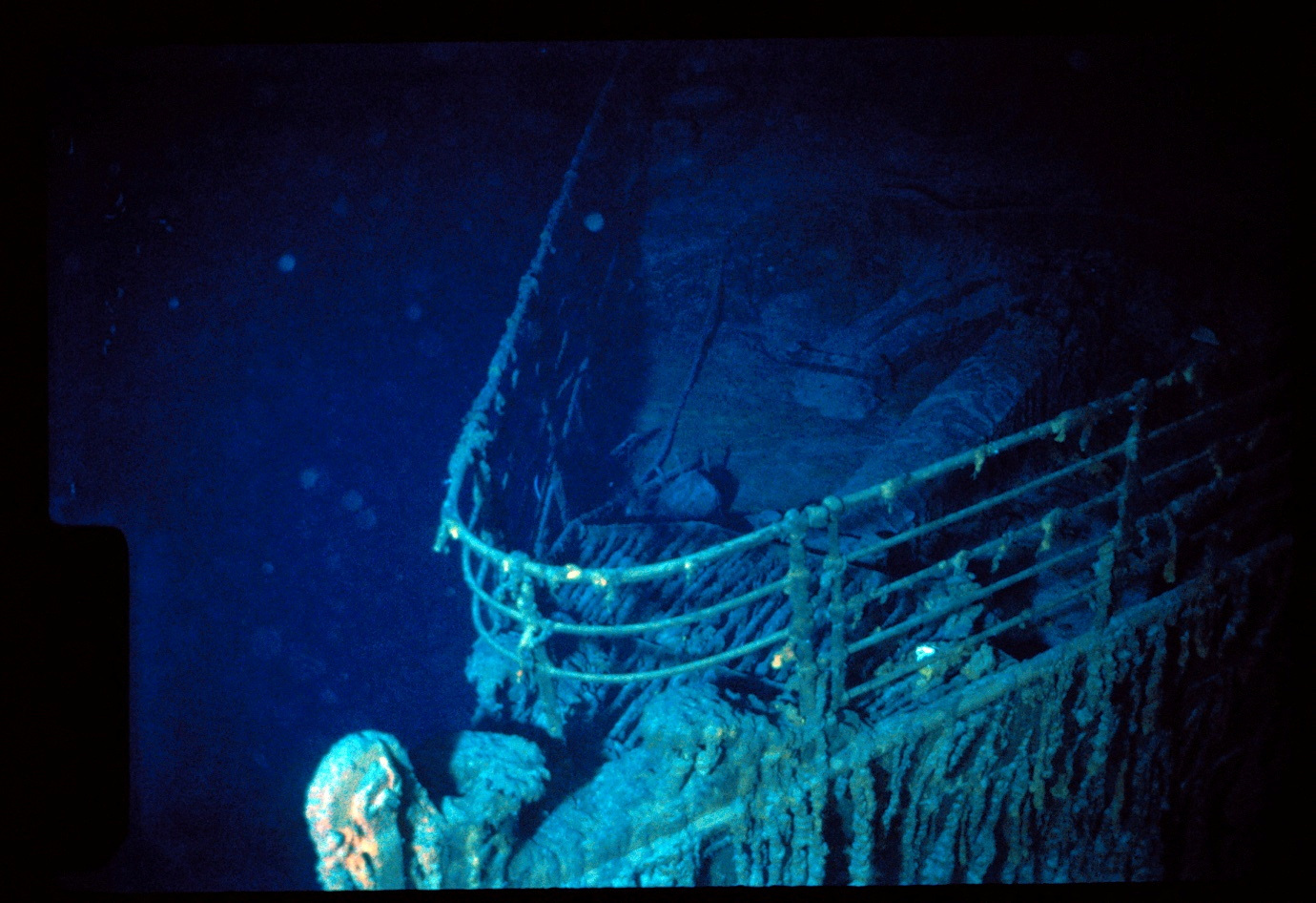 Bow of the Titanic wreck which looks an aqua colour with barnacles on it. The ocean water is a deep, dark blue