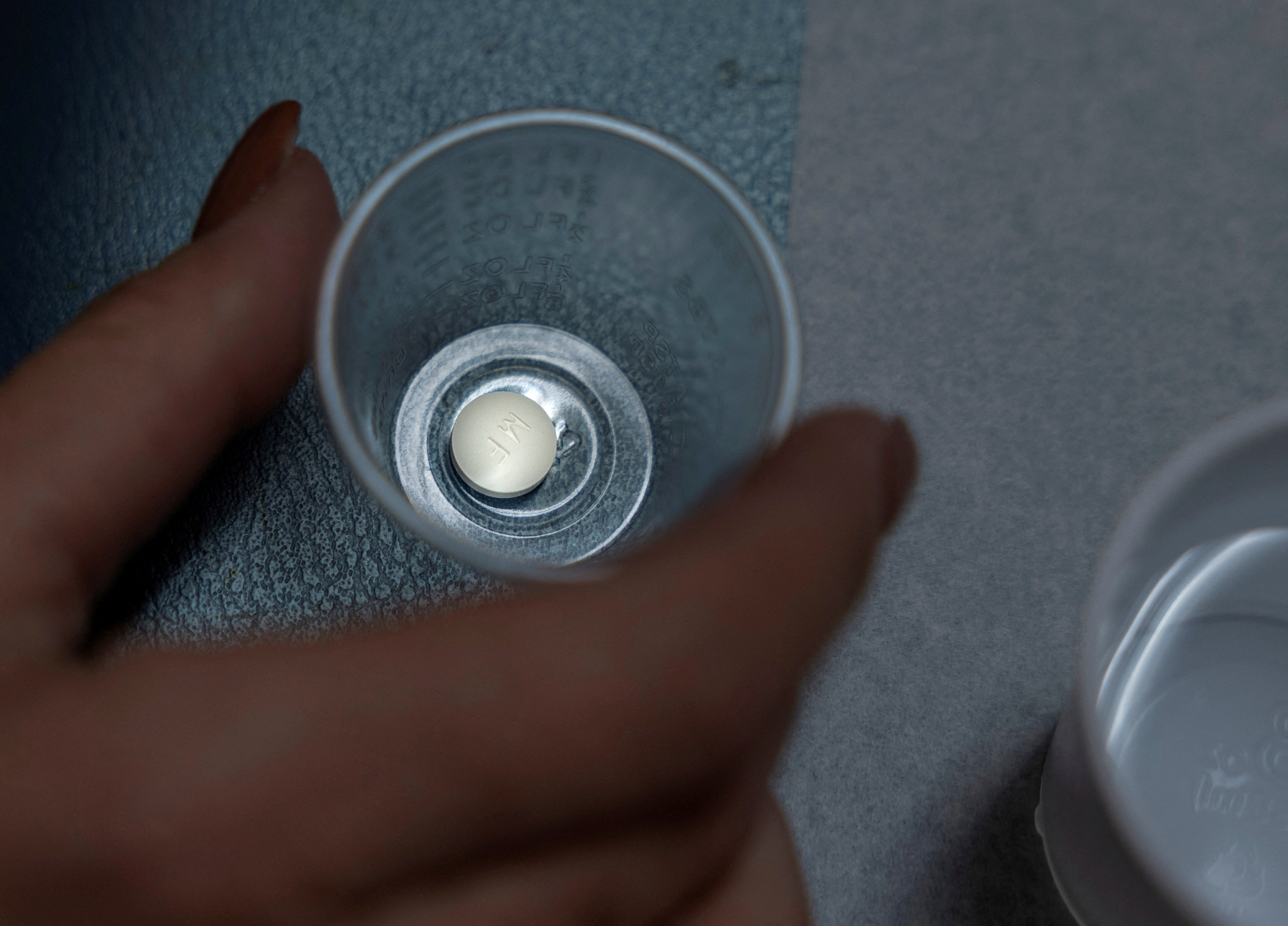 A manicured hand grabs a tiny plastic cup containing a round, white pill