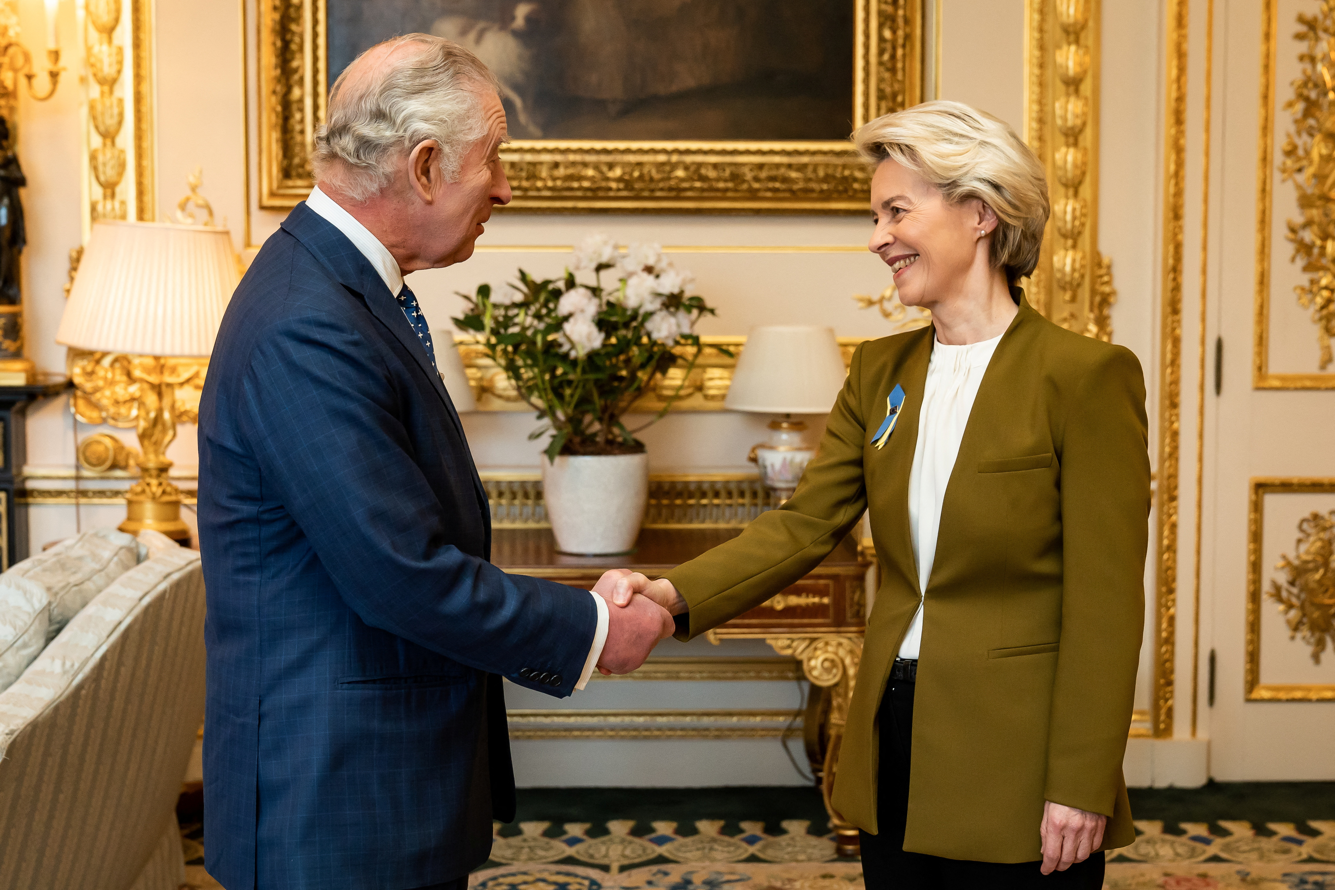 European Commission President Ursula von der Leyen meets King Charles III at Windsor Castle. The two are shaking hands and smiling. Von der Leyen has a ribbon in the blue and yellow of Ukraine on her lapel. The room has gilded walls and there is a plant with white flowers on the ornate table behind them. 