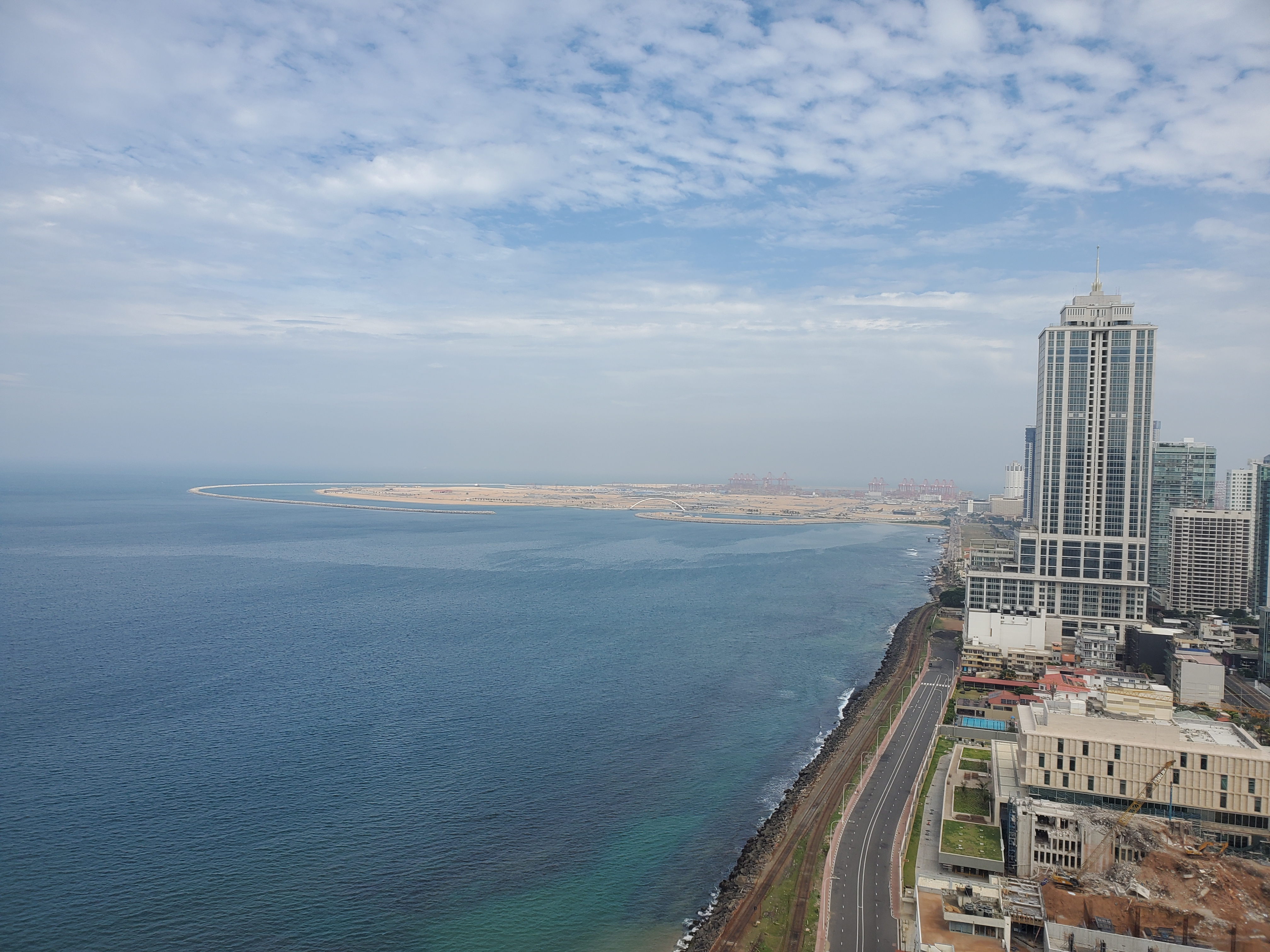 An aerial view shows the ocean on the left with Colombo city on the right, a road separating the two.