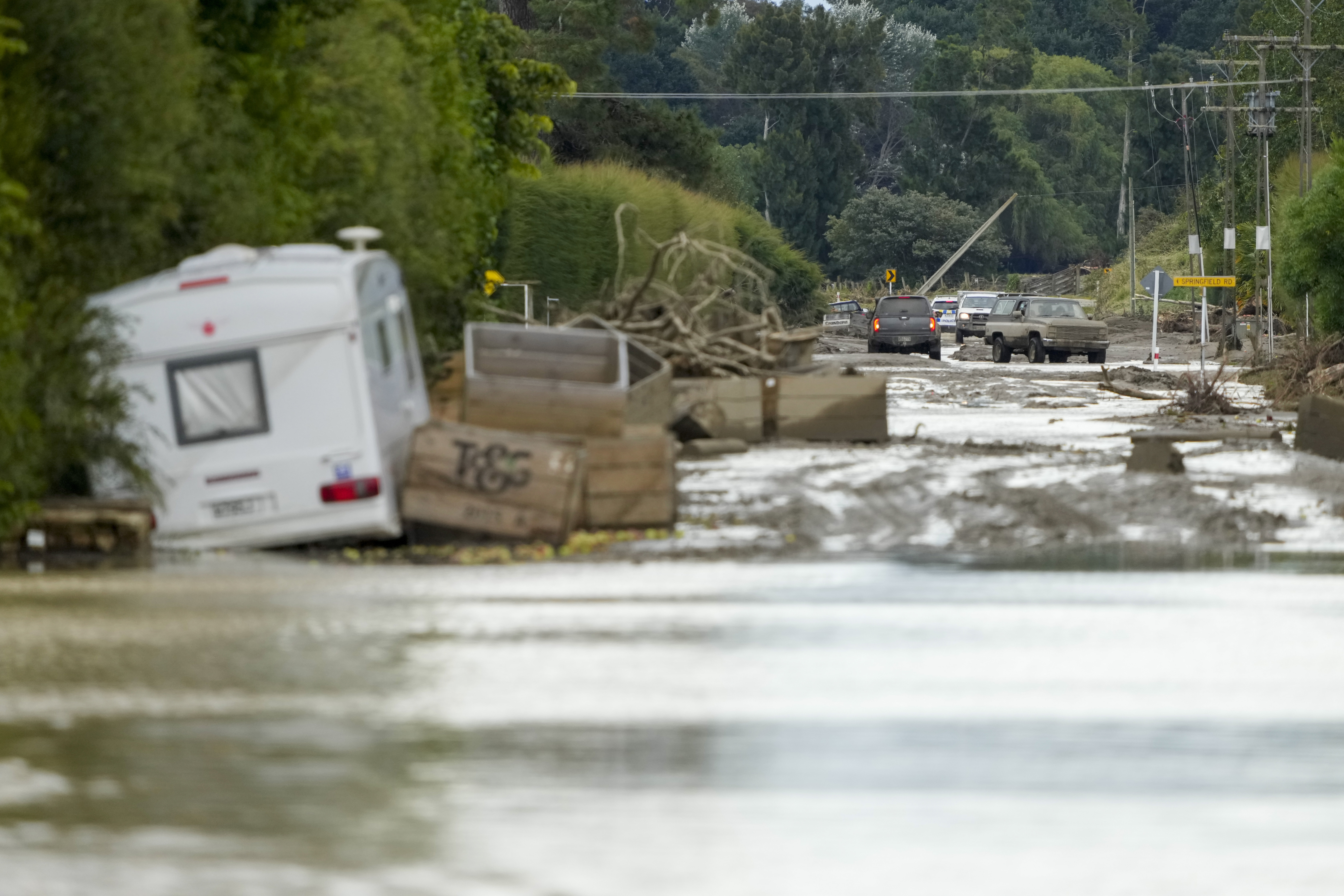 A caravan, tree debris and wooden crates in piles of mud on a road near Napier. There is flooding in the front of the photograph and mud covered trucks in the background
