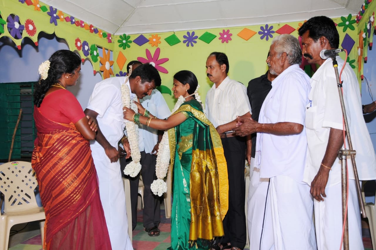 A photo of a group of people and a woman putting a scarf made of flowers around a man's neck.