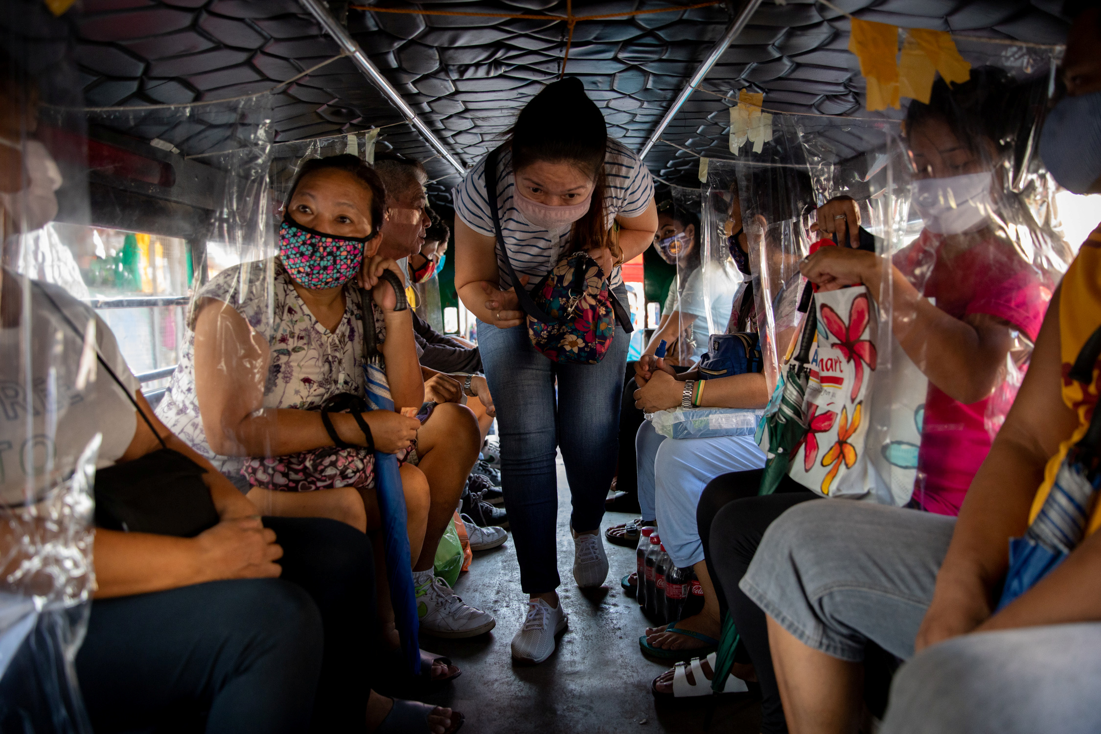 A woman in a face mask bending over as she moves down the central aisle of a jeepney to find a seat. There are passengers sitting on benches on either side of the vehicle.