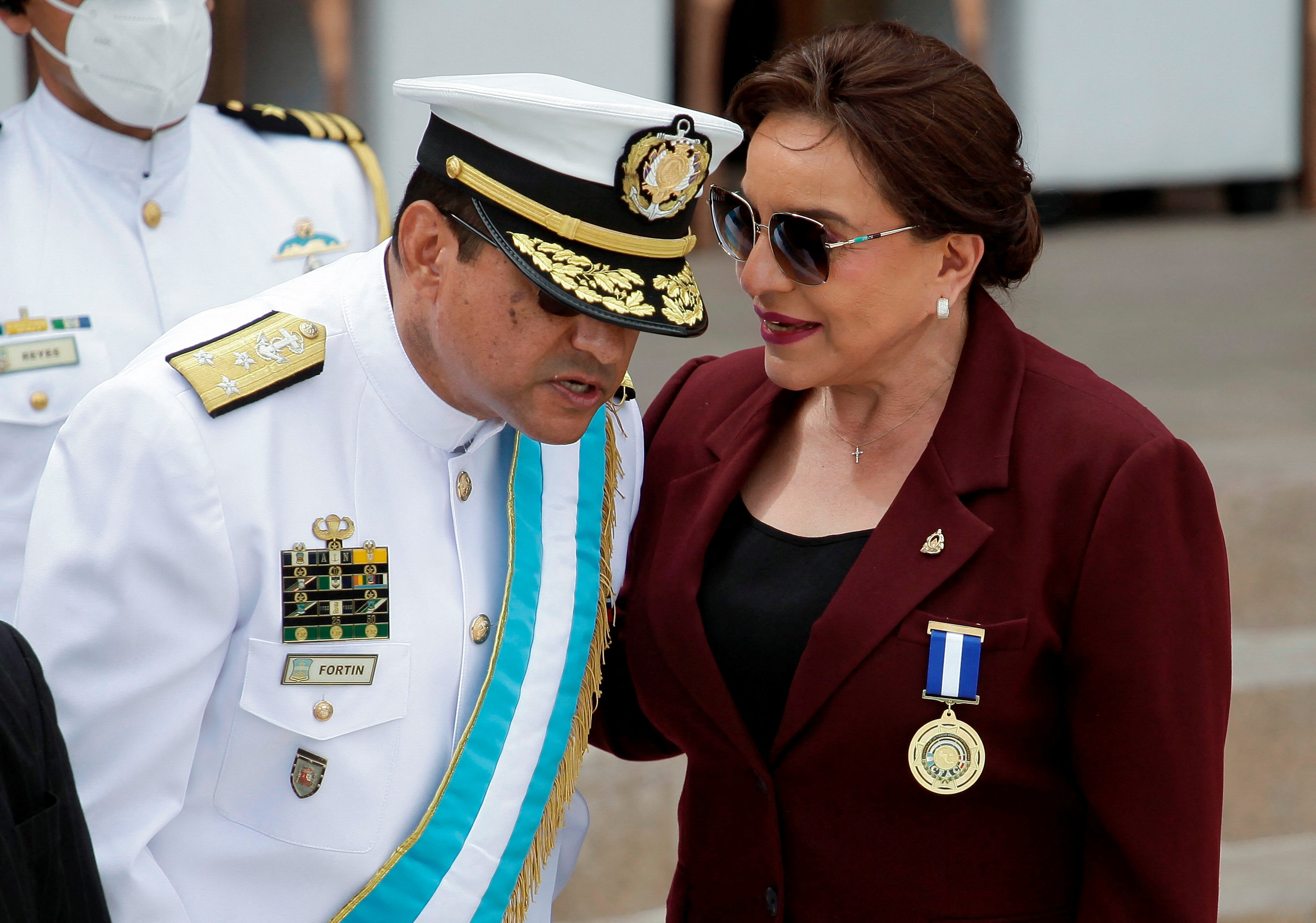 Honduras President Xiomara Castro talking to the Chairman of the Joint Chiefs of Staff at a military event. He is in ceremonial uniform and stooping slightly to hear her. She is wearing a burgundy jacket and sunglasses.
