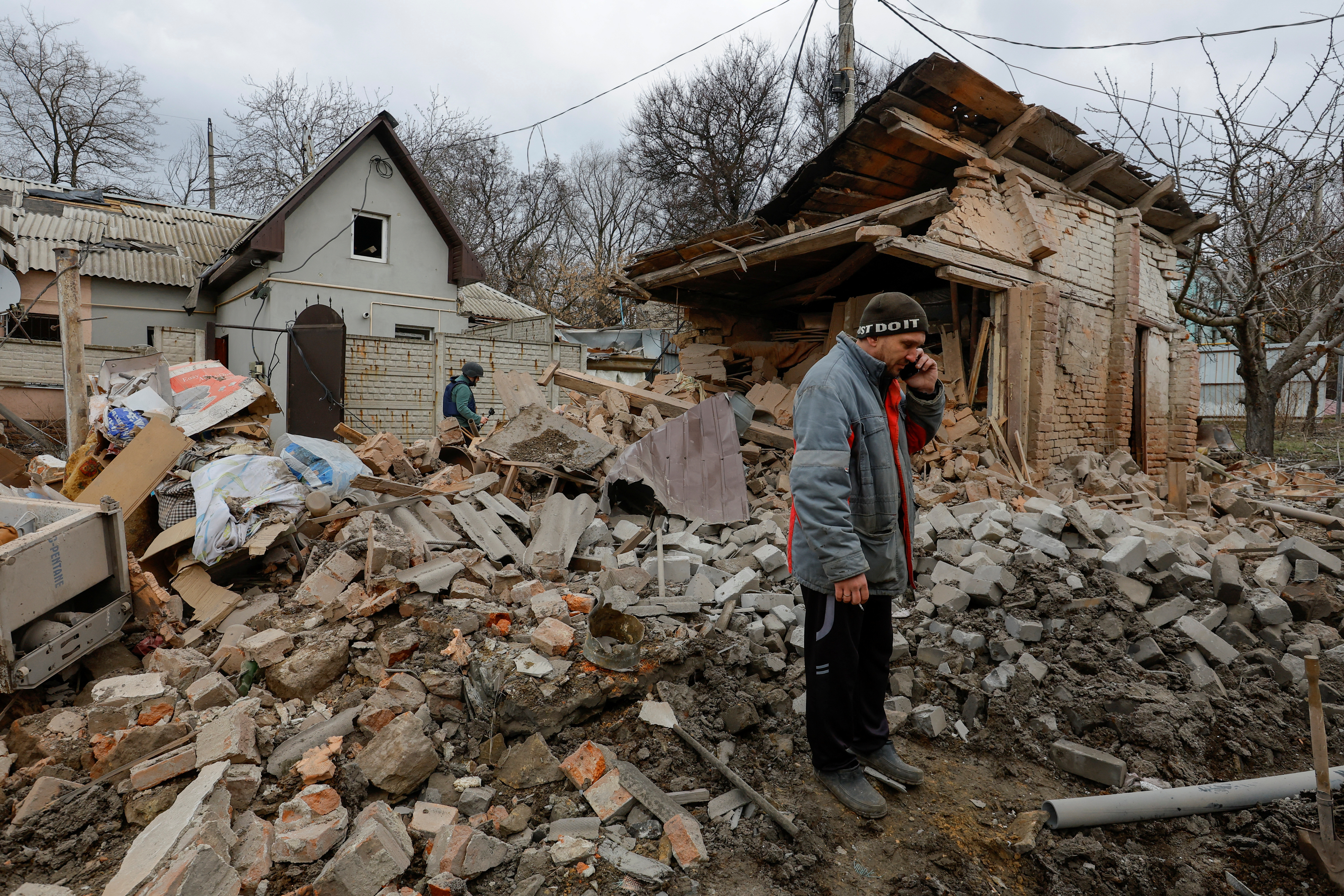 A man talks on the phone in front of damaged buildings. Debris is all around him.