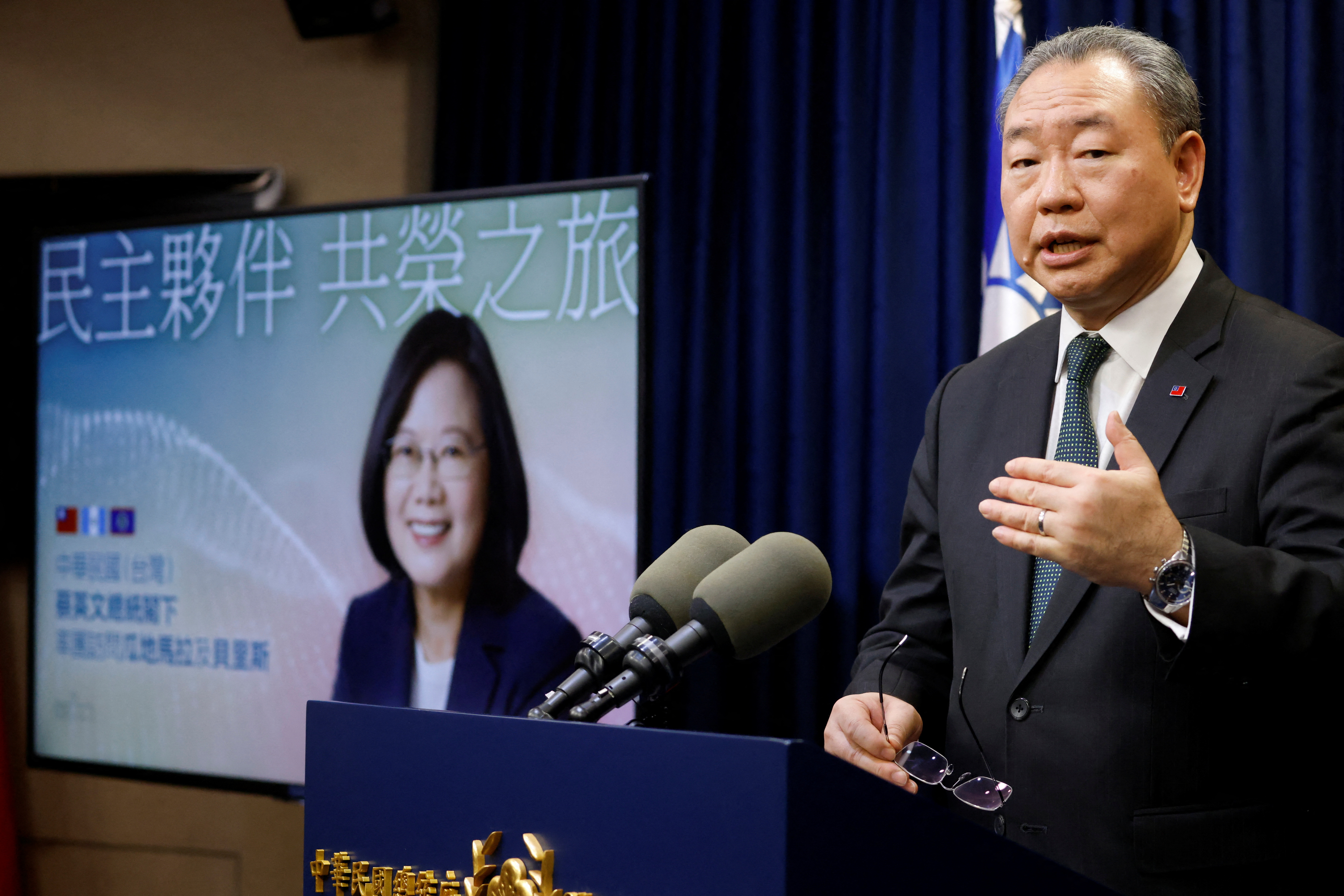 A man at a lectern speaks into two microphones, as a video screen next to him shows the image of Taiwan's president