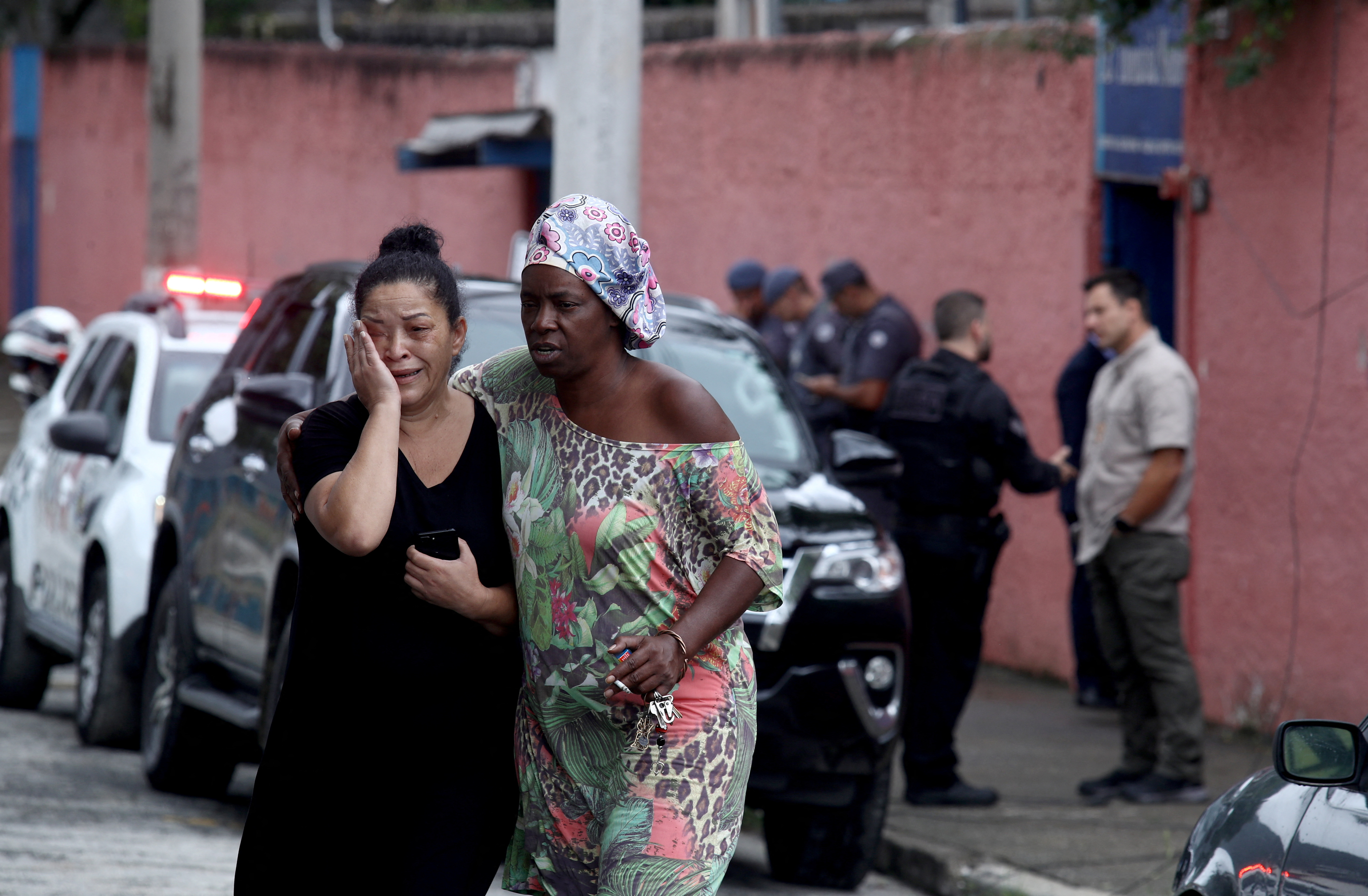 A woman wipes away tears while another wraps her arm around her on a street in front of a red wall. Police cars with flashing red lights can be seen in the background.