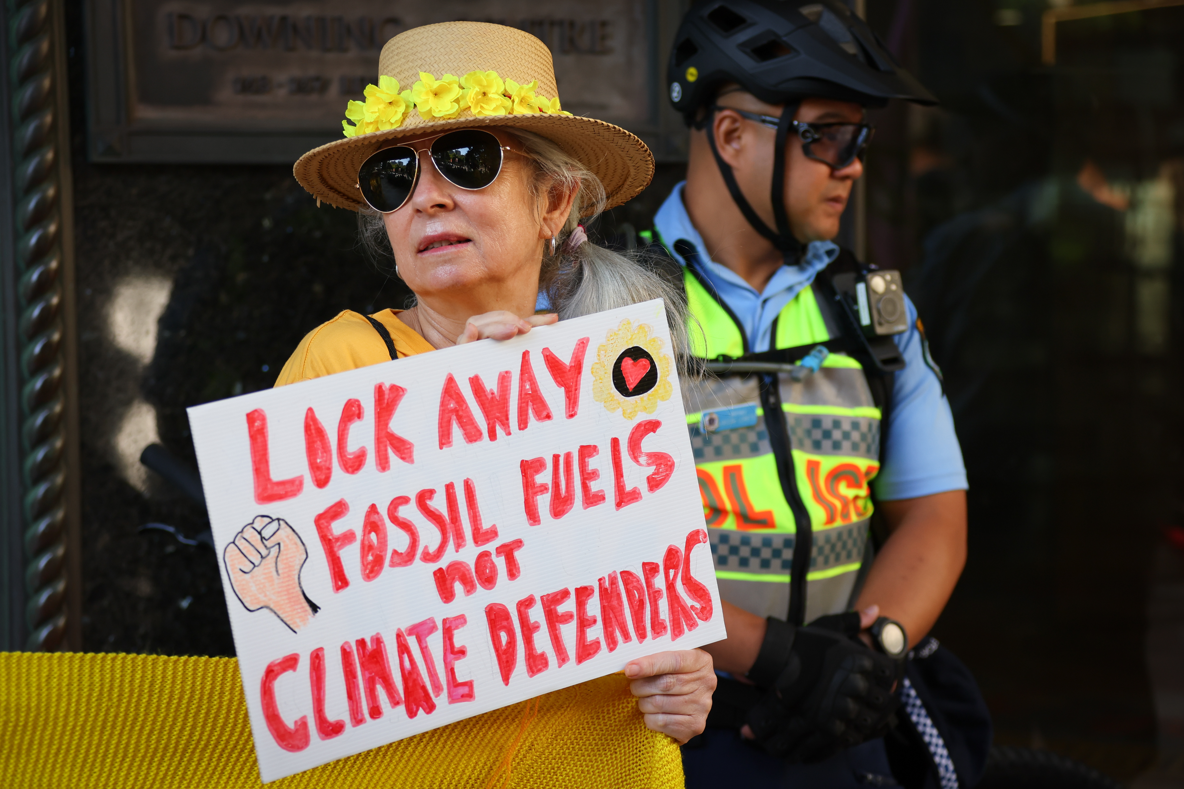 A woman holding a sign in support of Deanna 'Violet' CoCo. It reads 'Lock away fossil fuels not climate defenders'. The woman is wearing sunglasses and a straw hat with yellow flowers around the brim. There is a policeman in the background.