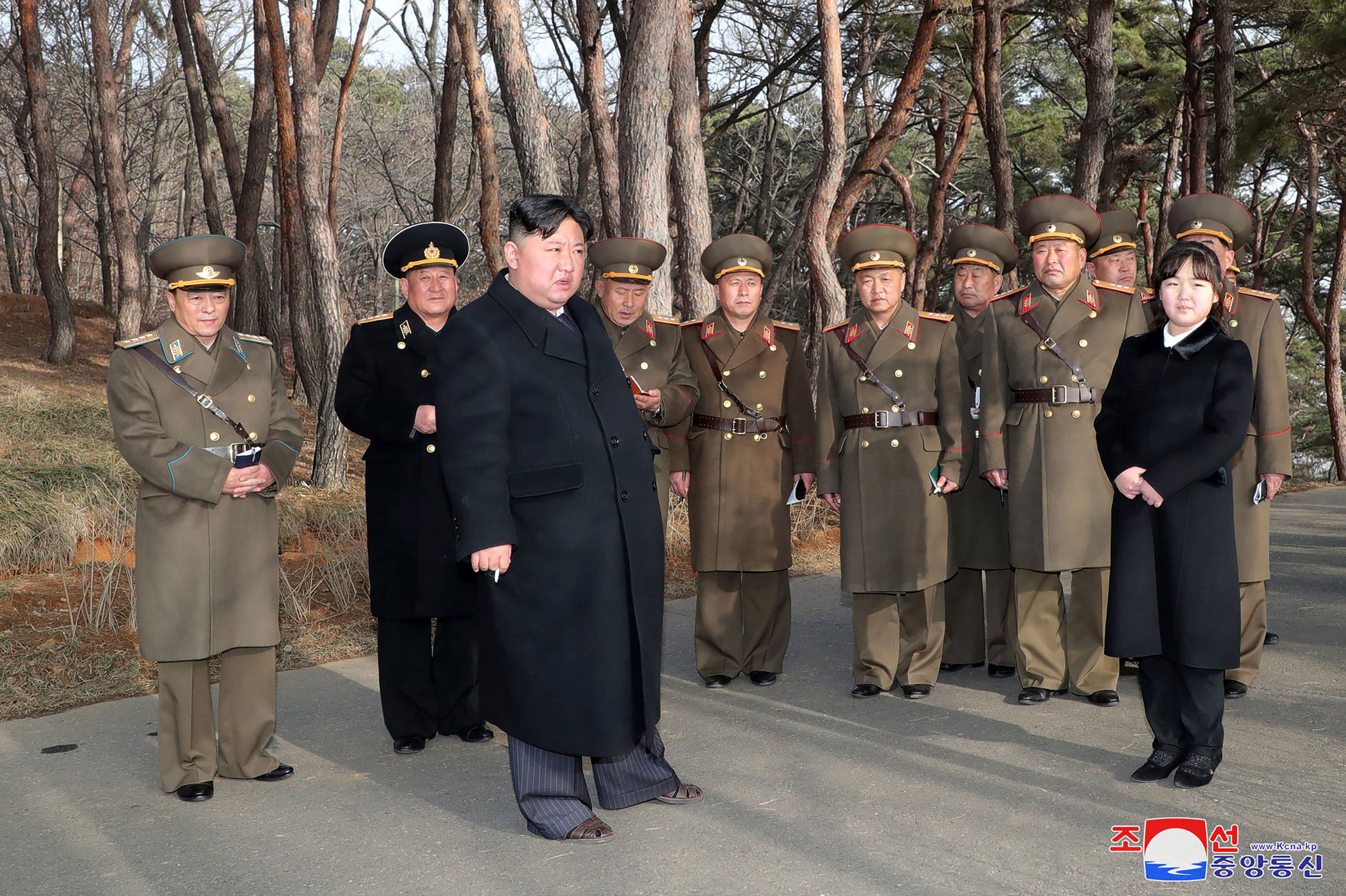 Kim Jong Un and his daughter standing outside. There are military officials in uniform behind them. Kim and his daughter are both wearing black.