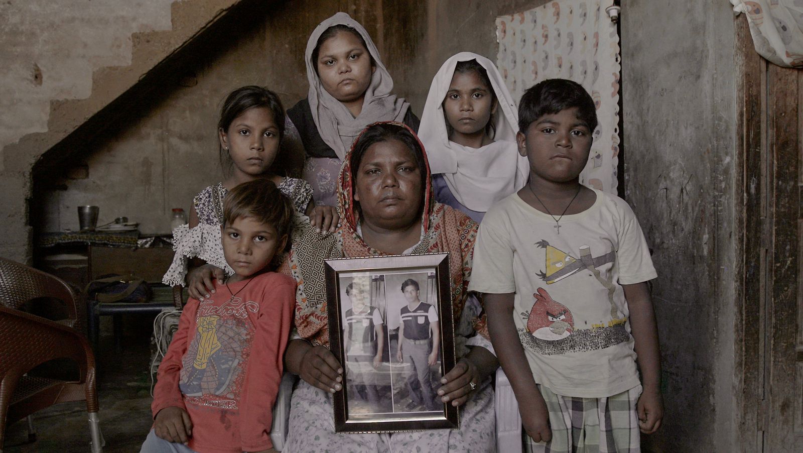 A photo of a woman sitting and holding a frame with two identical photos of a man side by side with five people standing around her.