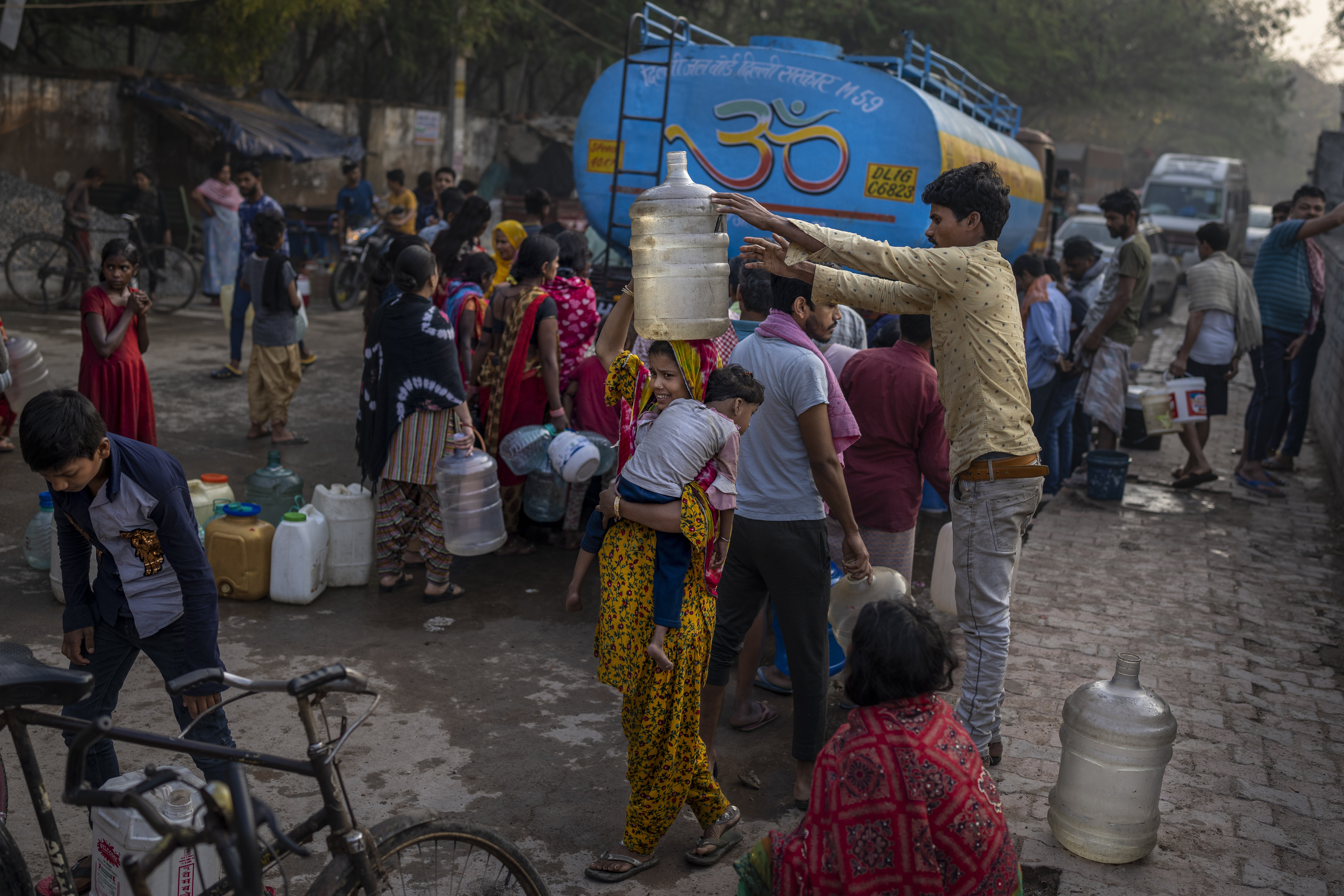 A woman balances a water can on her head while carrying a child as people collect water from a mobile water tanker on World Water Day in a residential area in New Delhi, India
