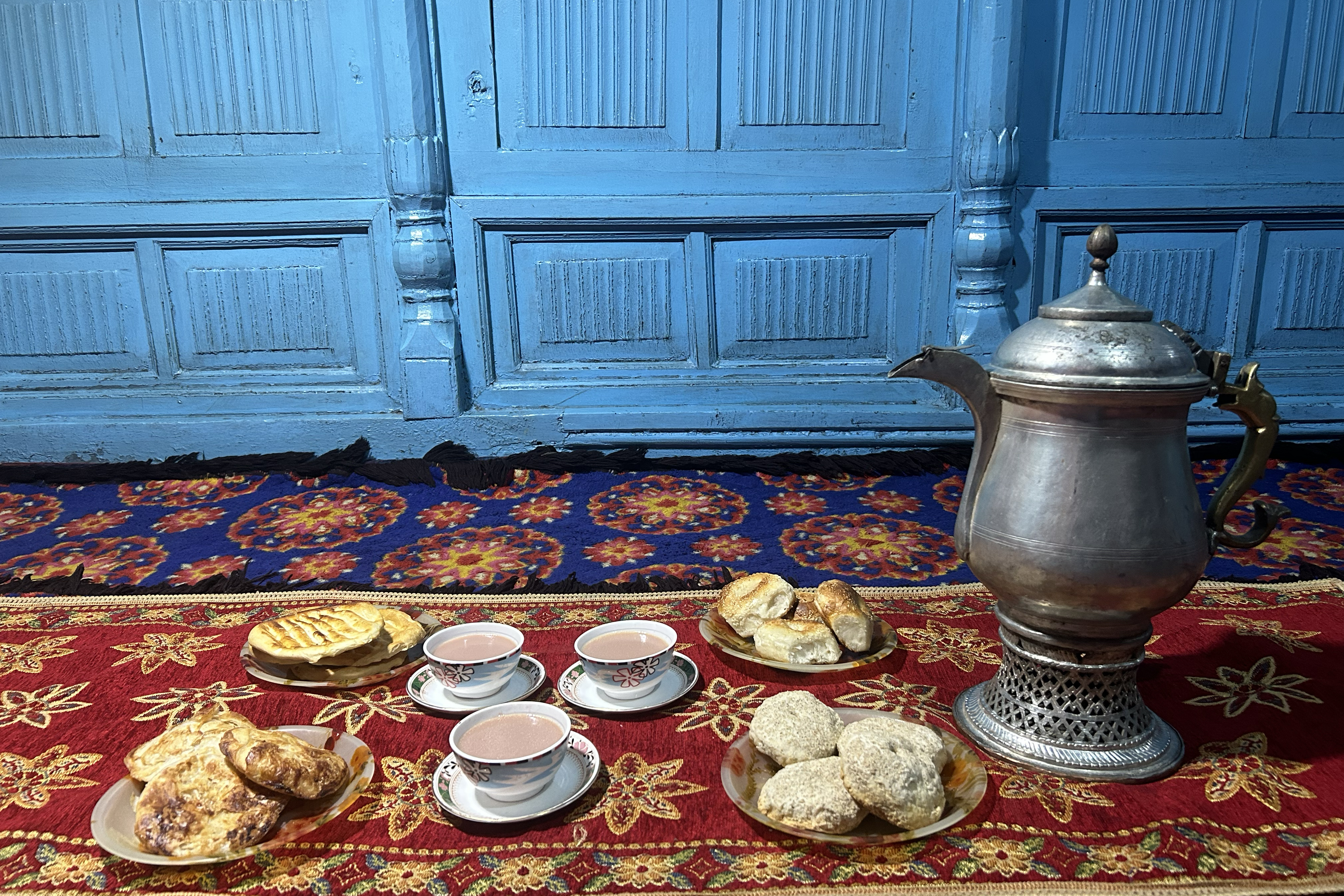 A samovar, three cups of noon chai and four plates of snacks laid out on a colourful carpet in front of a turqoise wooden wall