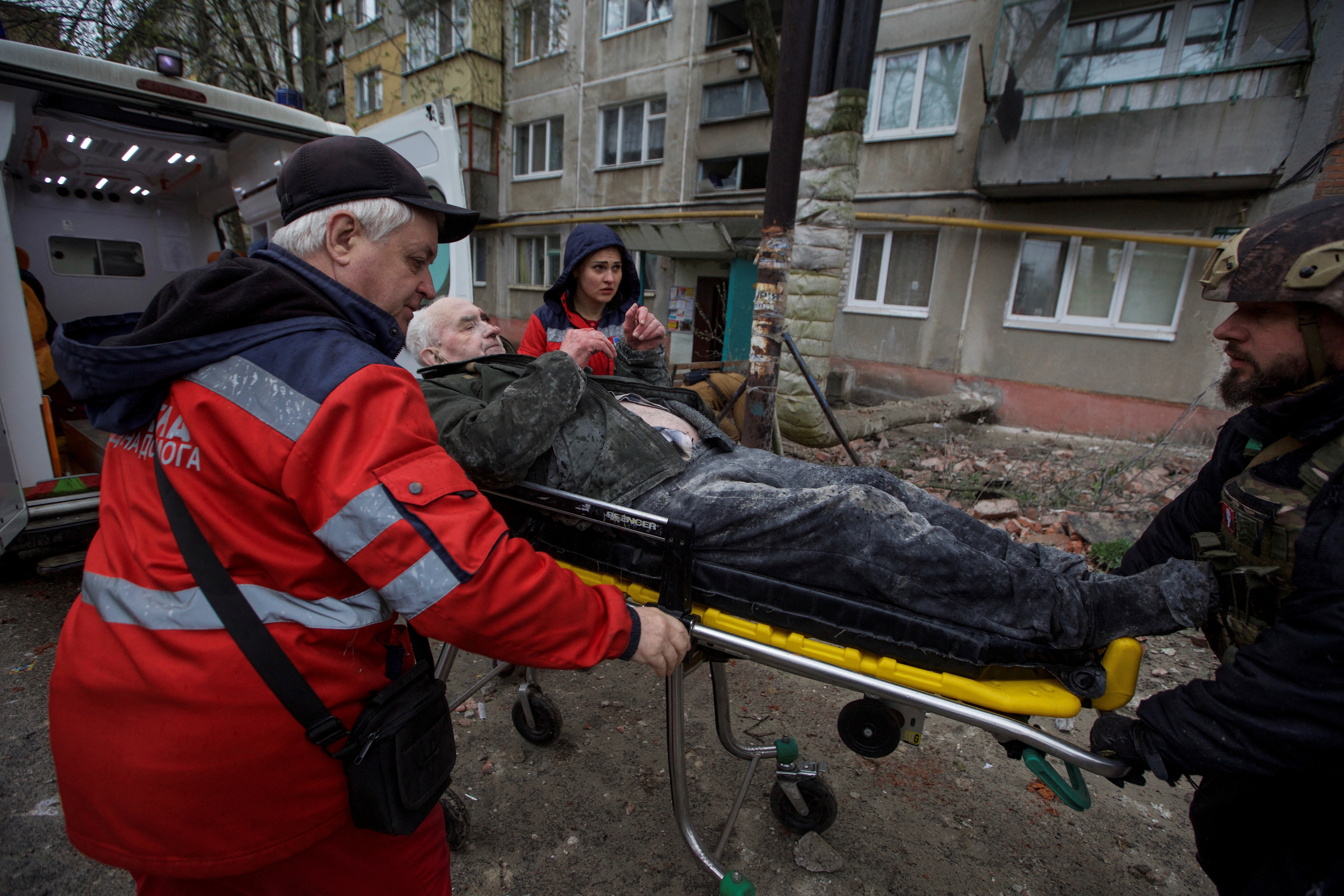 An older man being stretchered to an ambulance after an apartment building in Sloviansk was shelled. There is an emegency worker on each side and a soldier at the end by his feet. The damaged building is behind.