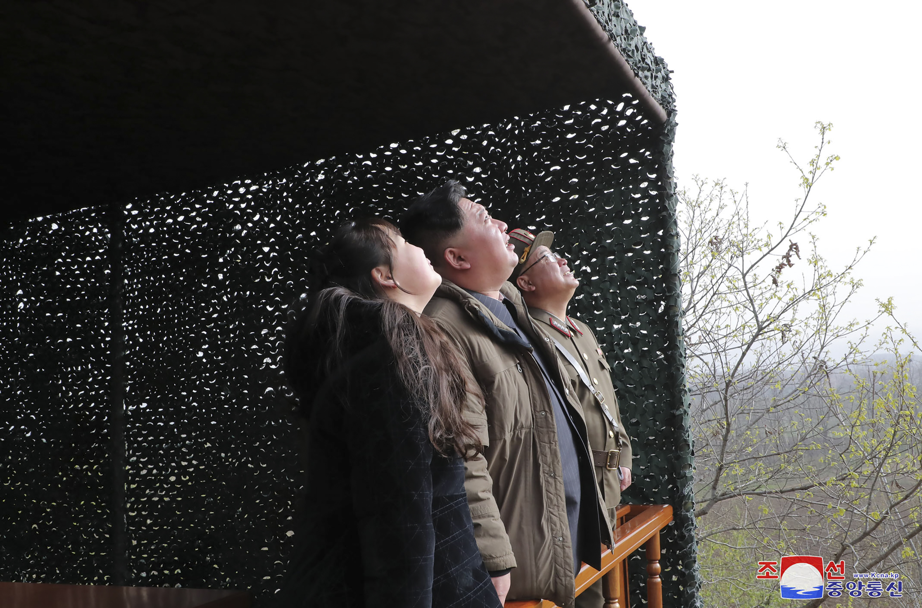 Kim Jong Un and his daughter looking upwards as the missile takes off. They are standing side by side in a tent. here is a military official beside Kim Kong Un.