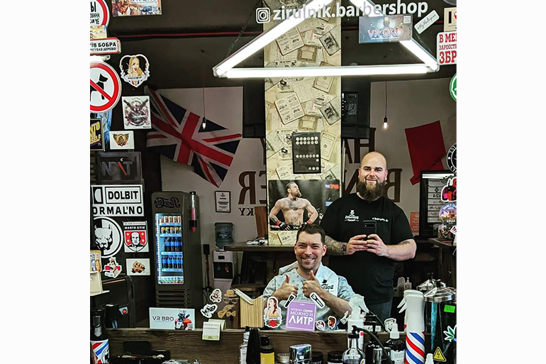 A photo of Igor on the right and Vitalii on the left looking at themselves through a mirror. Igor is sitting on a chair and Vitalii is standing behind him.