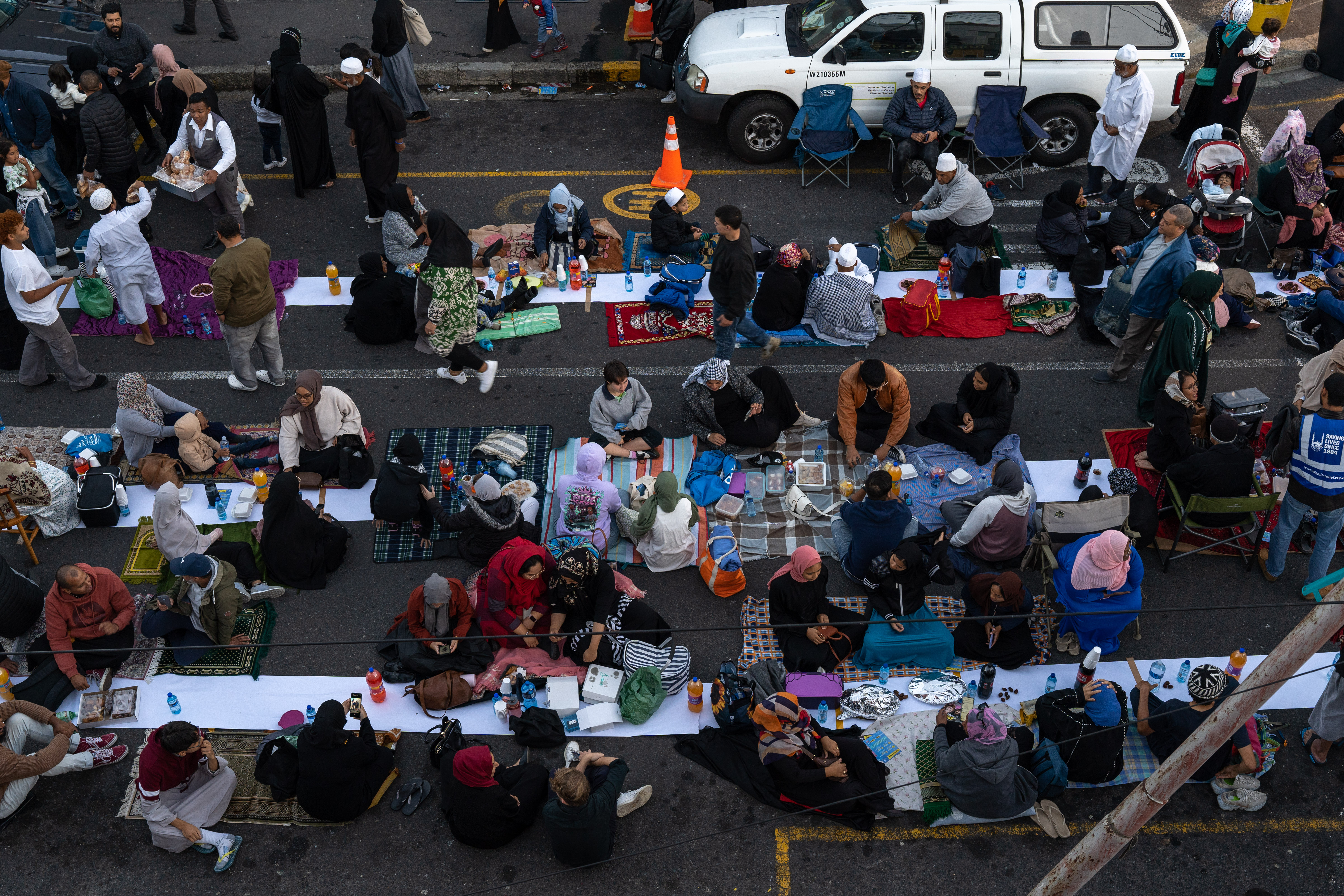 An aerial view of groups of people sitting around chairs, tables and blankets, eating and drinking.
