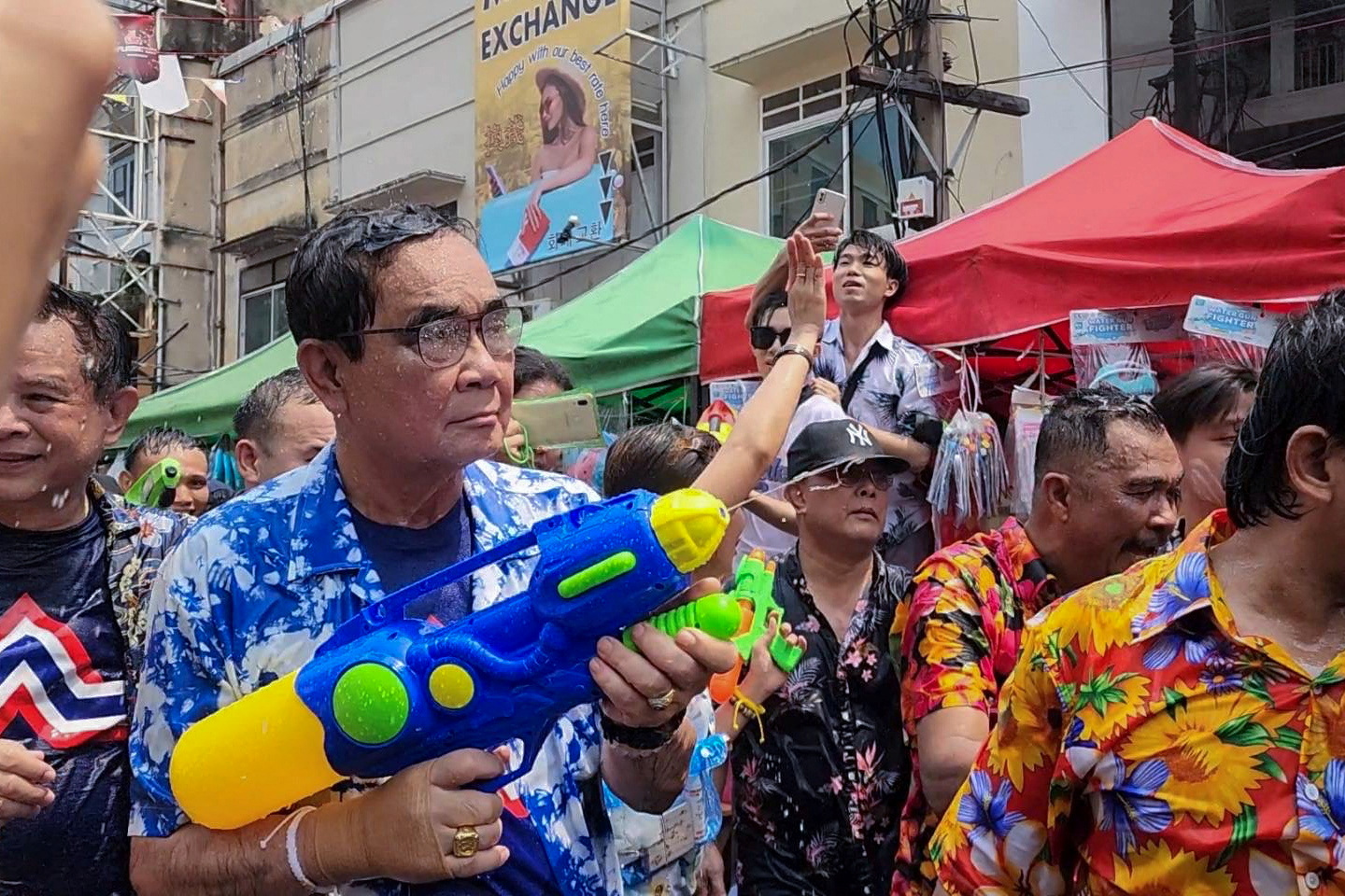 Prayuth Chan-ocha in a blue Hawaiian shirt at Songkran festivities in Bangkok. He has a large blue water gun, Others are spraying him with water and his hair is wet. He is not smiling.