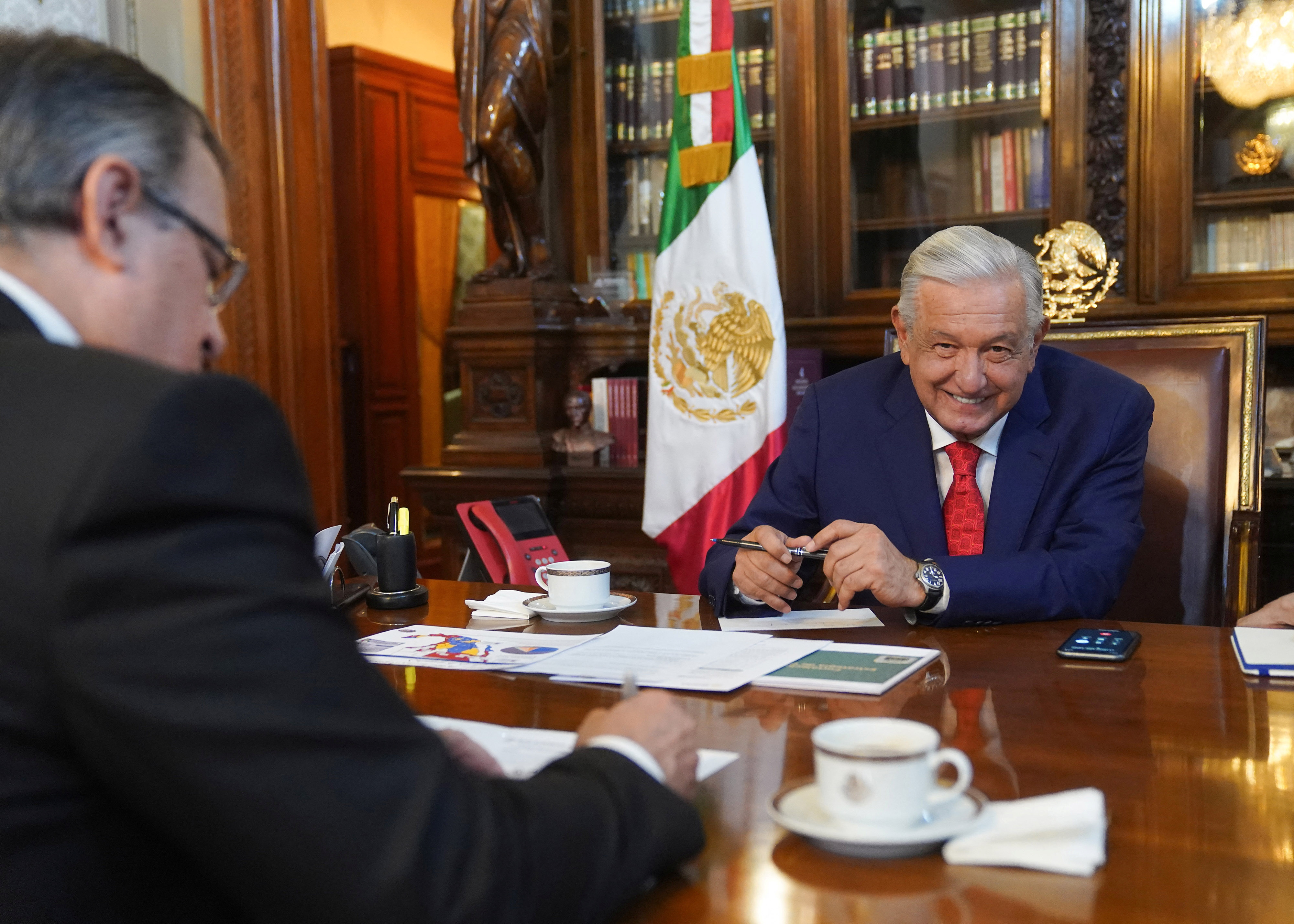 Mexico's President Andres Manuel Lopez Obrador and Mexico's Foreign Secretary Marcelo Ebrard hold a call at the National Palace in Mexico City. Lopez Obrador is seen on the far side of the wooden table, in front of a Mexican flag and a glass-shielded book case.